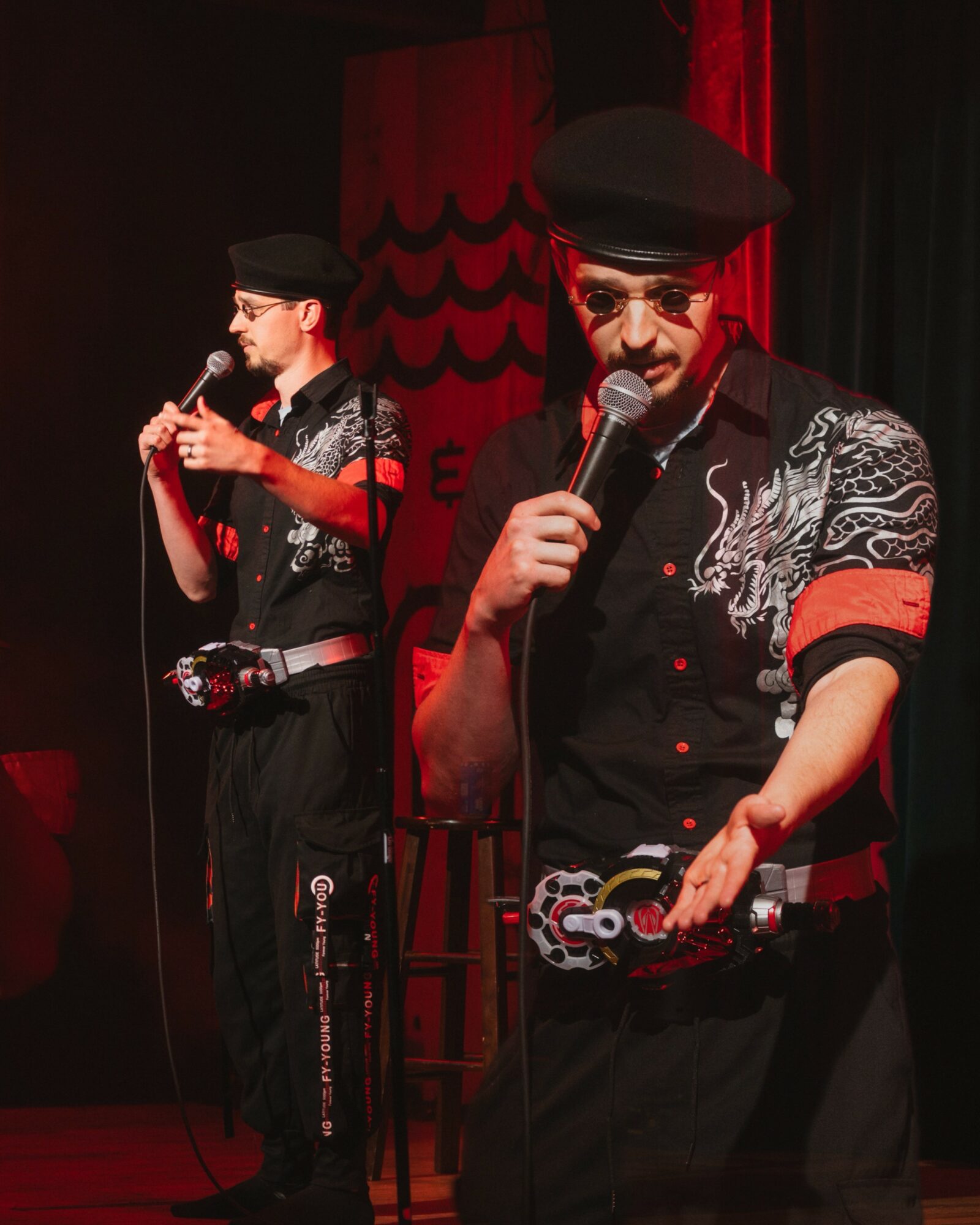 Two performers on stage, one holding a microphone and the other playing a wind instrument, both wearing black outfits with embroidery, hats, and sunglasses, illuminated by red stage lighting.