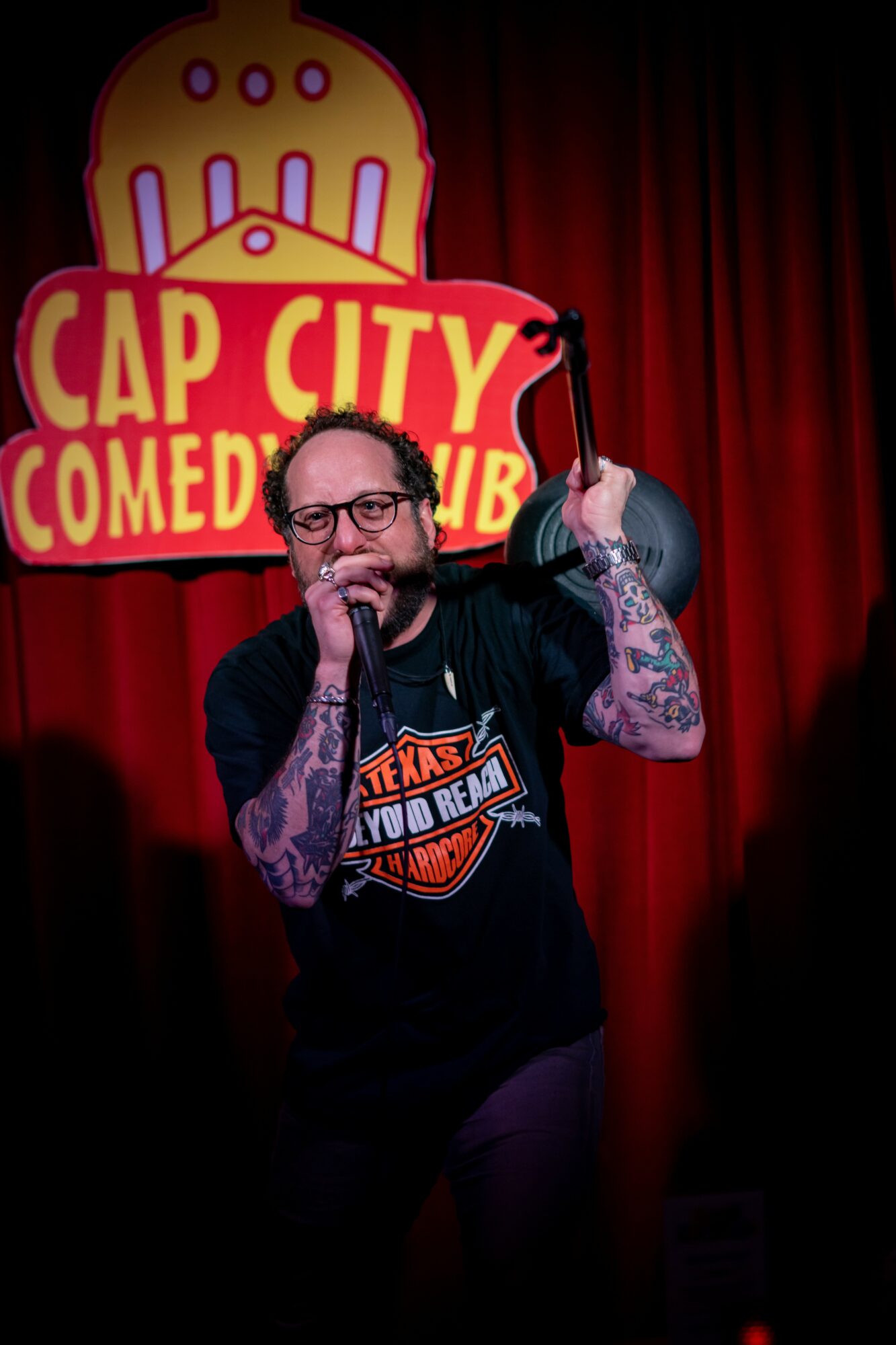 Comedian performing on stage with a microphone, red curtain background, and a sign reading 'Cap City Comedy Club'.