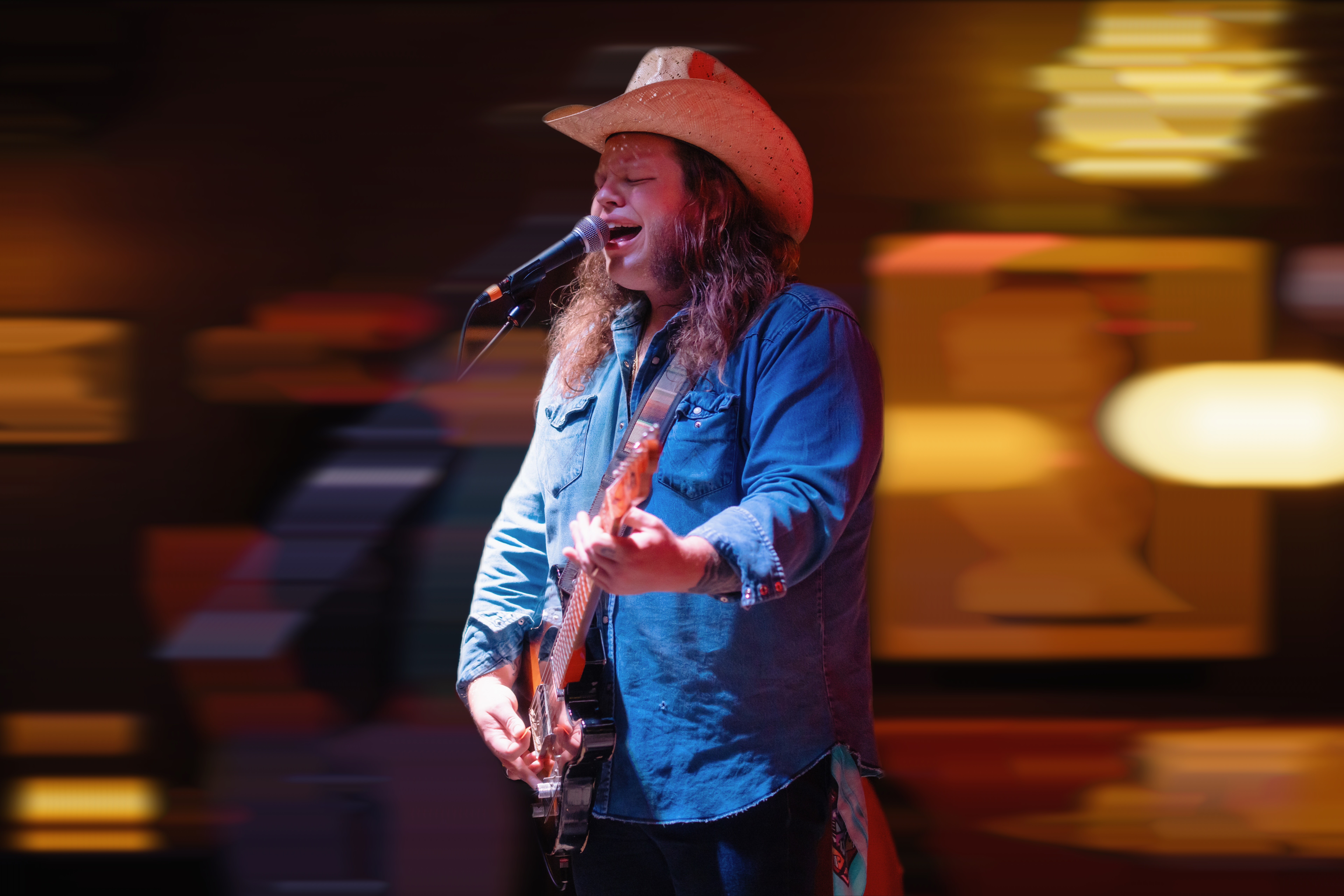 Person wearing a cowboy hat playing guitar and singing on stage with blurred background lights.
