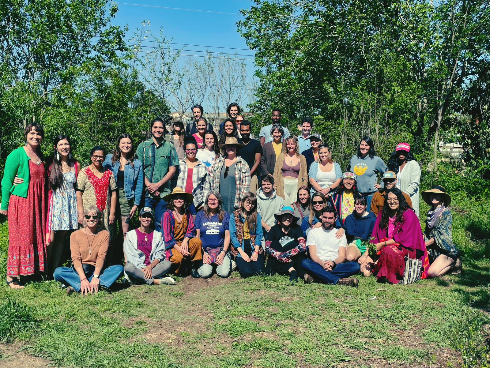 Group of people outdoors on grass with trees in background, posing for a photo, some sitting and some standing.
