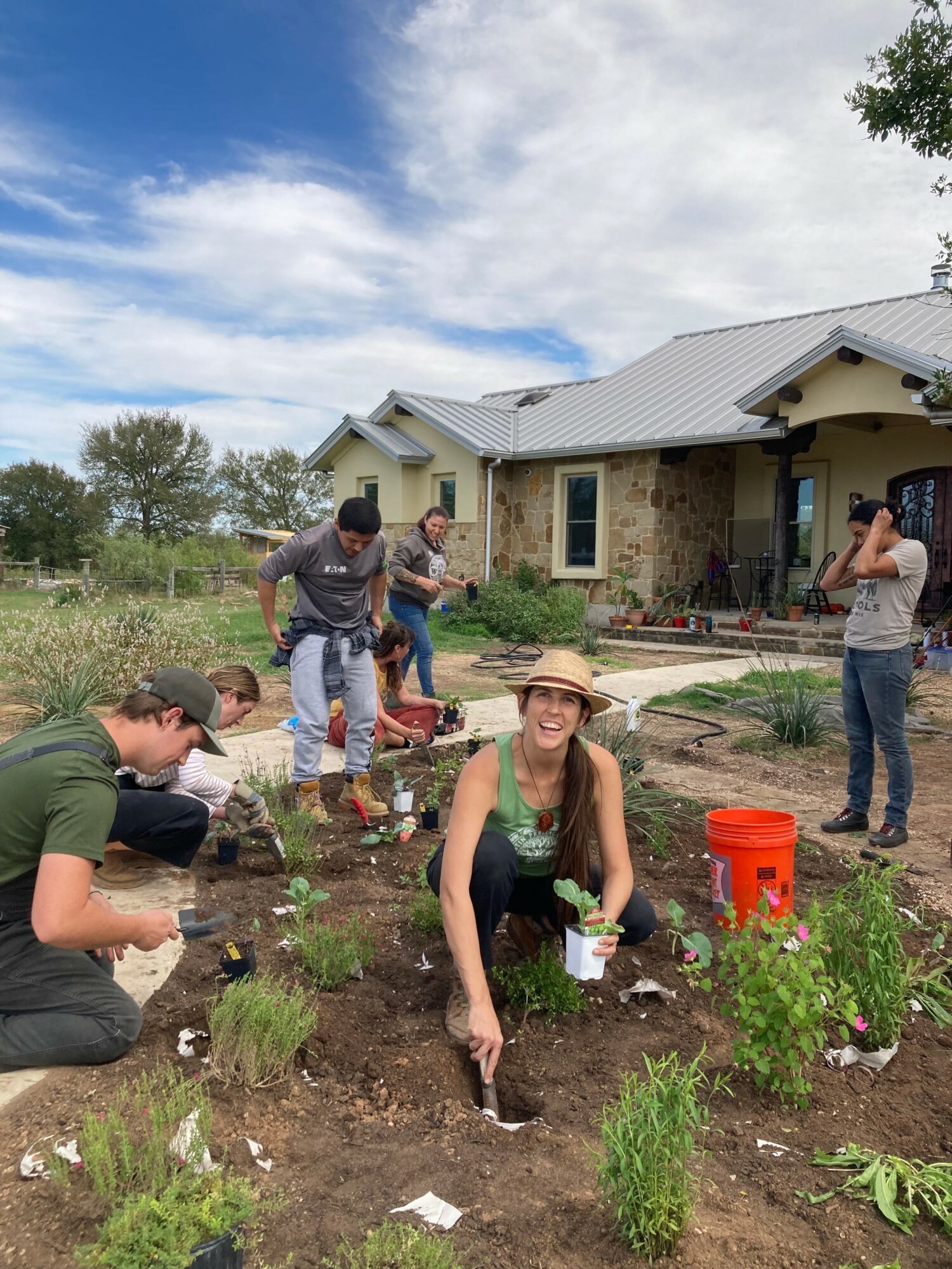 Group of people gardening in front of a house with a stone facade, some planting, and a woman smiling wearing a hat.