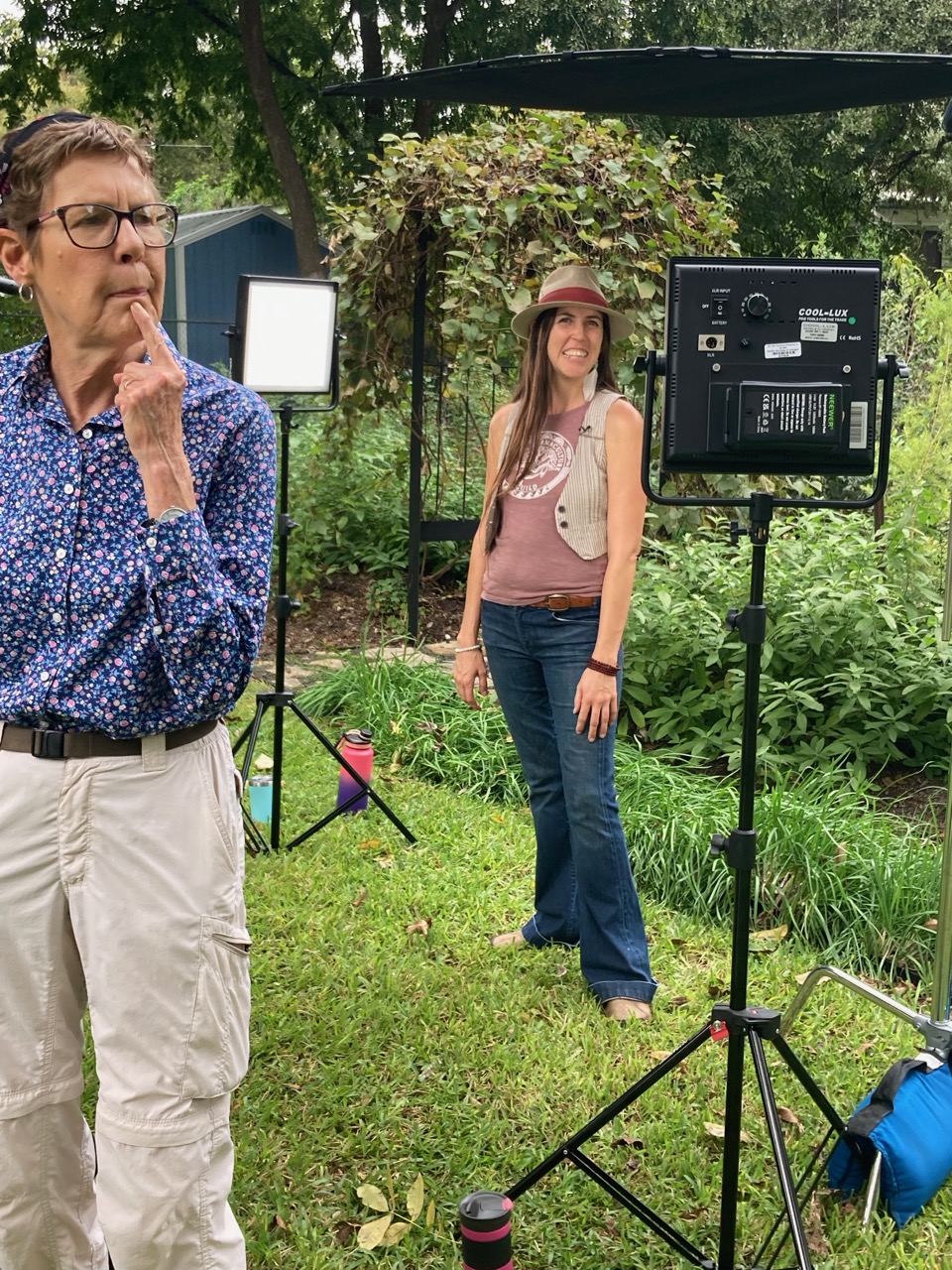 Two women outdoors with greenery, one in foreground with glasses and thoughtful expression, another in background smiling, standing near a camera setup.