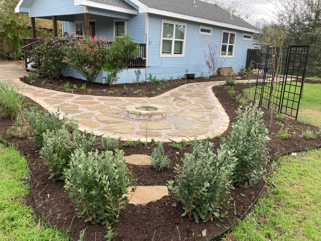 Stone pathway curves around a garden with bushes, leading to a blue house with a porch and garden bed.