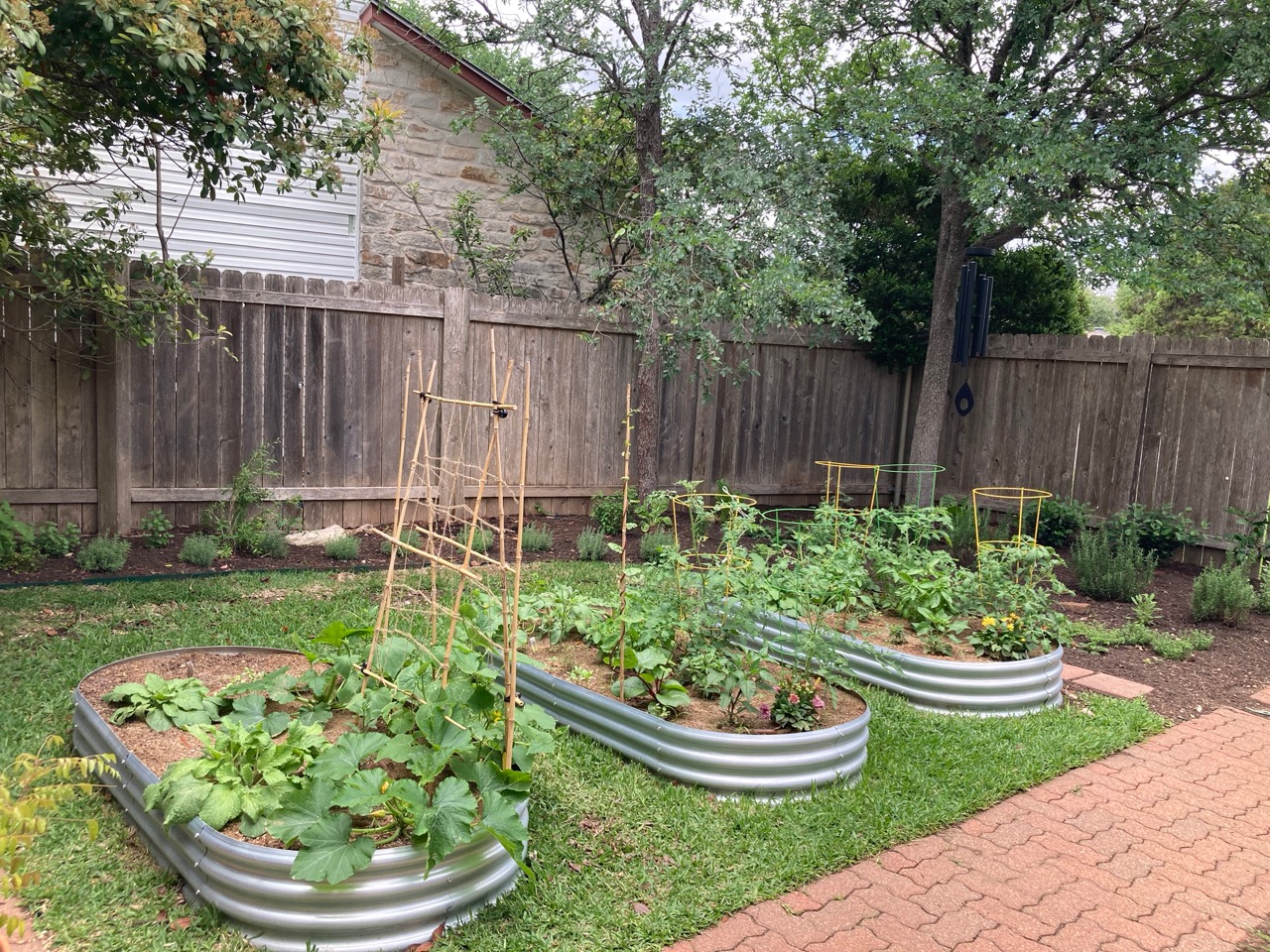 Three raised garden beds with plants, surrounded by a brick pathway and a wooden fence, with trees in the background.