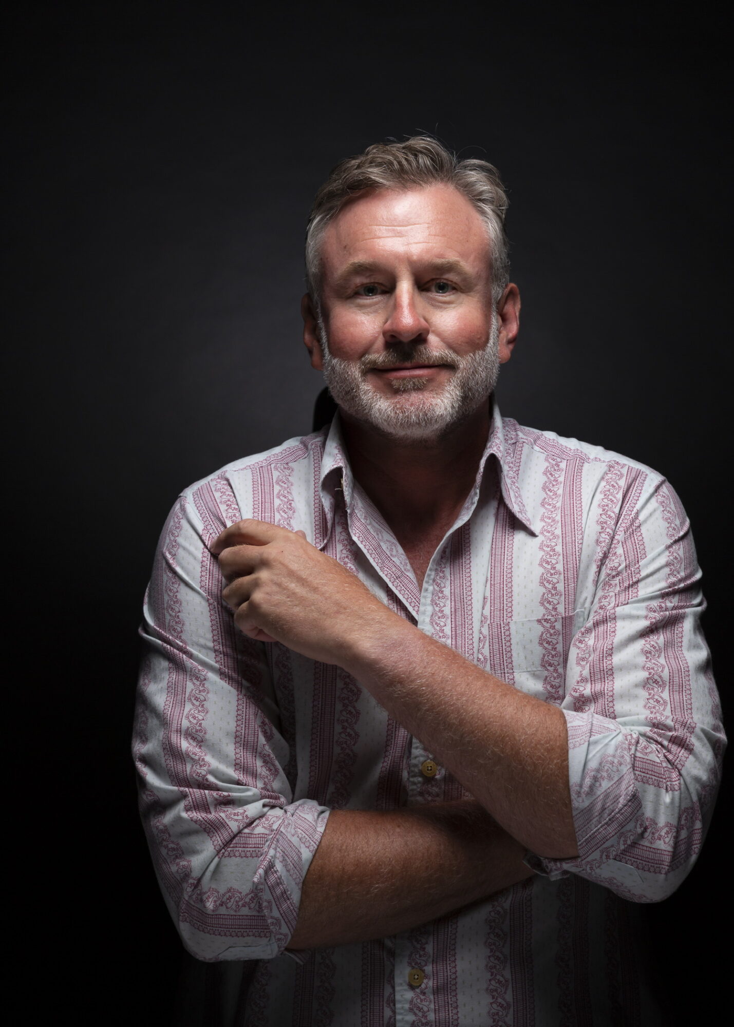 Portrait of a middle-aged man with gray hair and beard, wearing a striped shirt, smiling, with arms crossed, against a dark background.