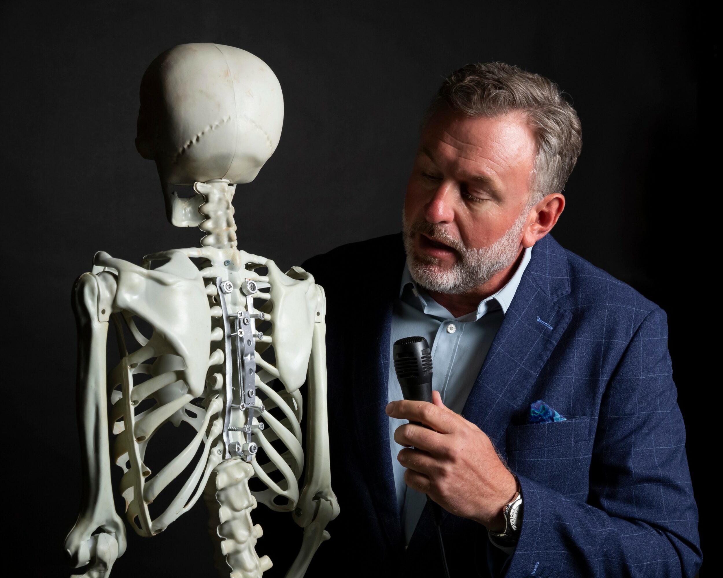 Man with gray hair and beard speaking into a microphone next to a human skeleton model against a dark background.