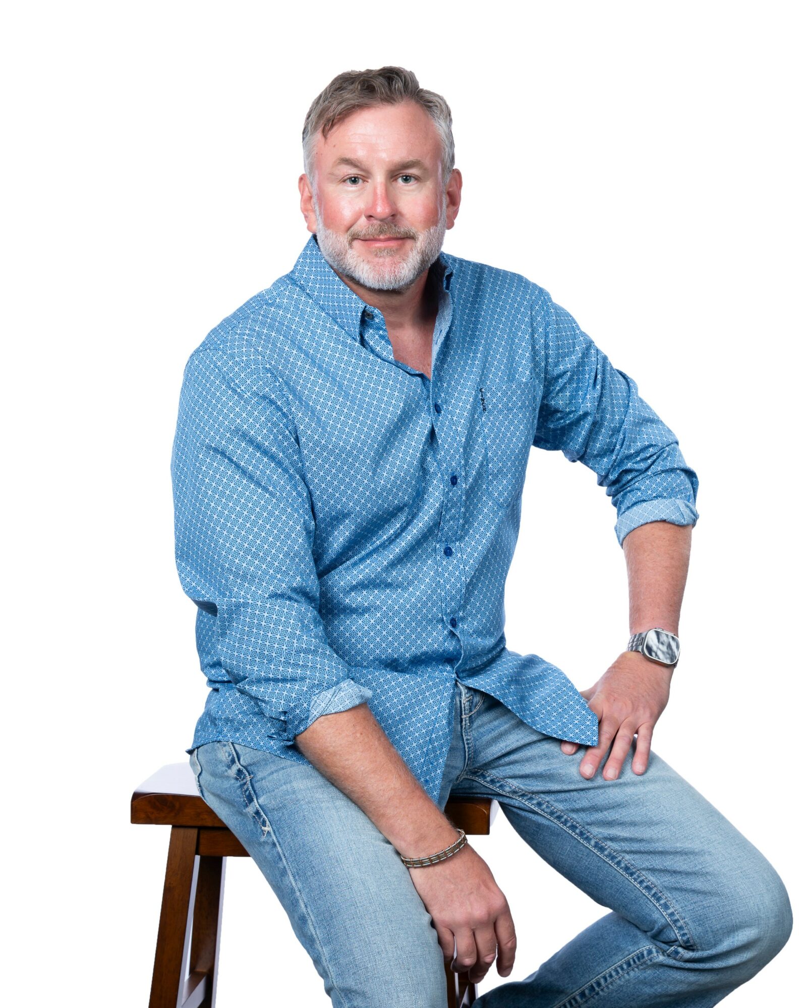 A middle-aged man with gray hair and beard, wearing a blue shirt and jeans, sitting on a wooden stool against a white background.