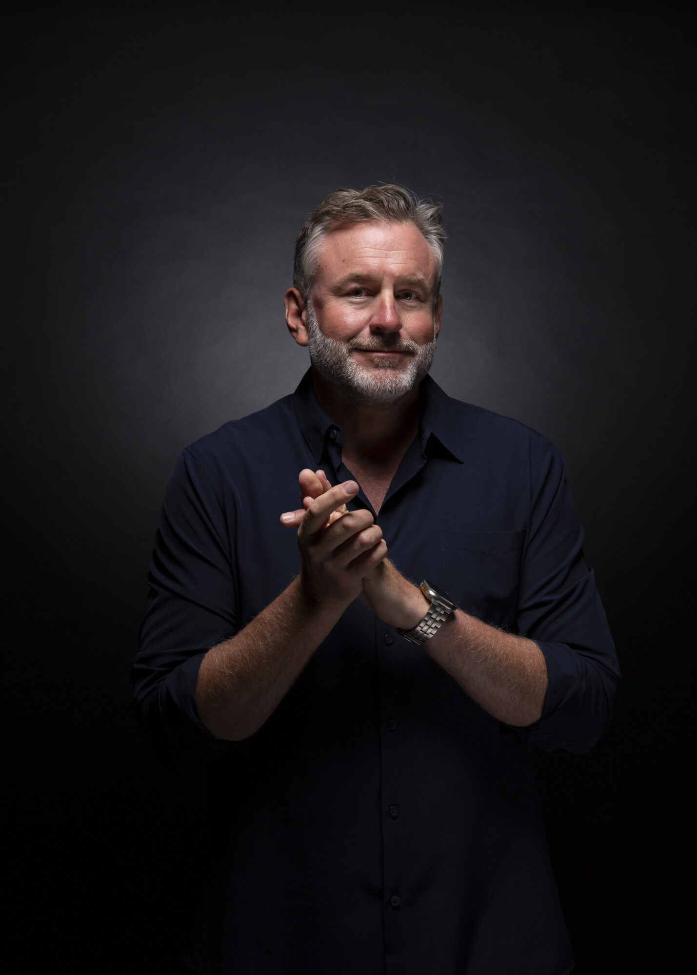 Man with gray hair and beard claps hands, wearing a dark shirt, against a dark background.