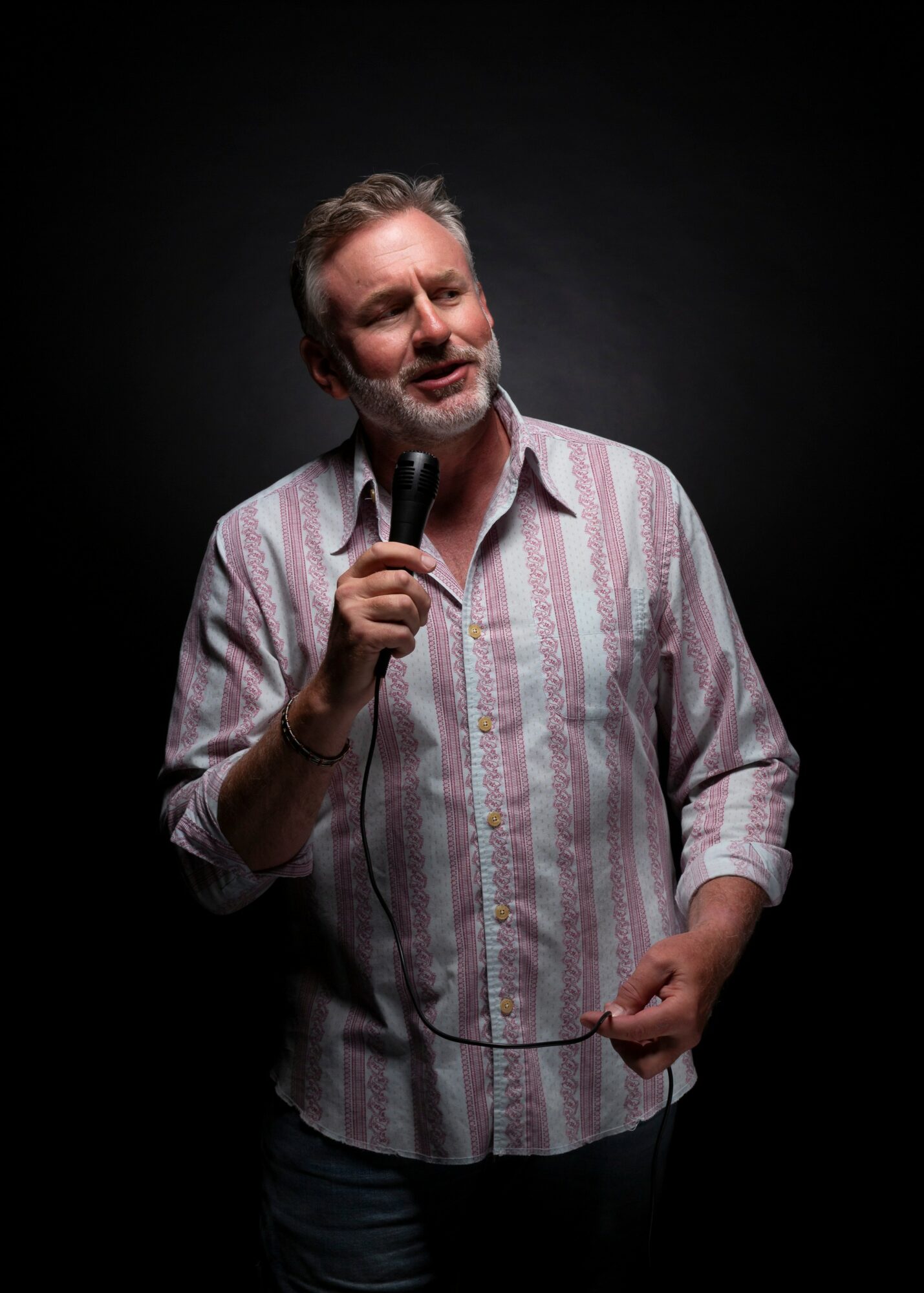 Man with gray hair and beard holding a microphone, wearing a striped shirt, against a dark background.