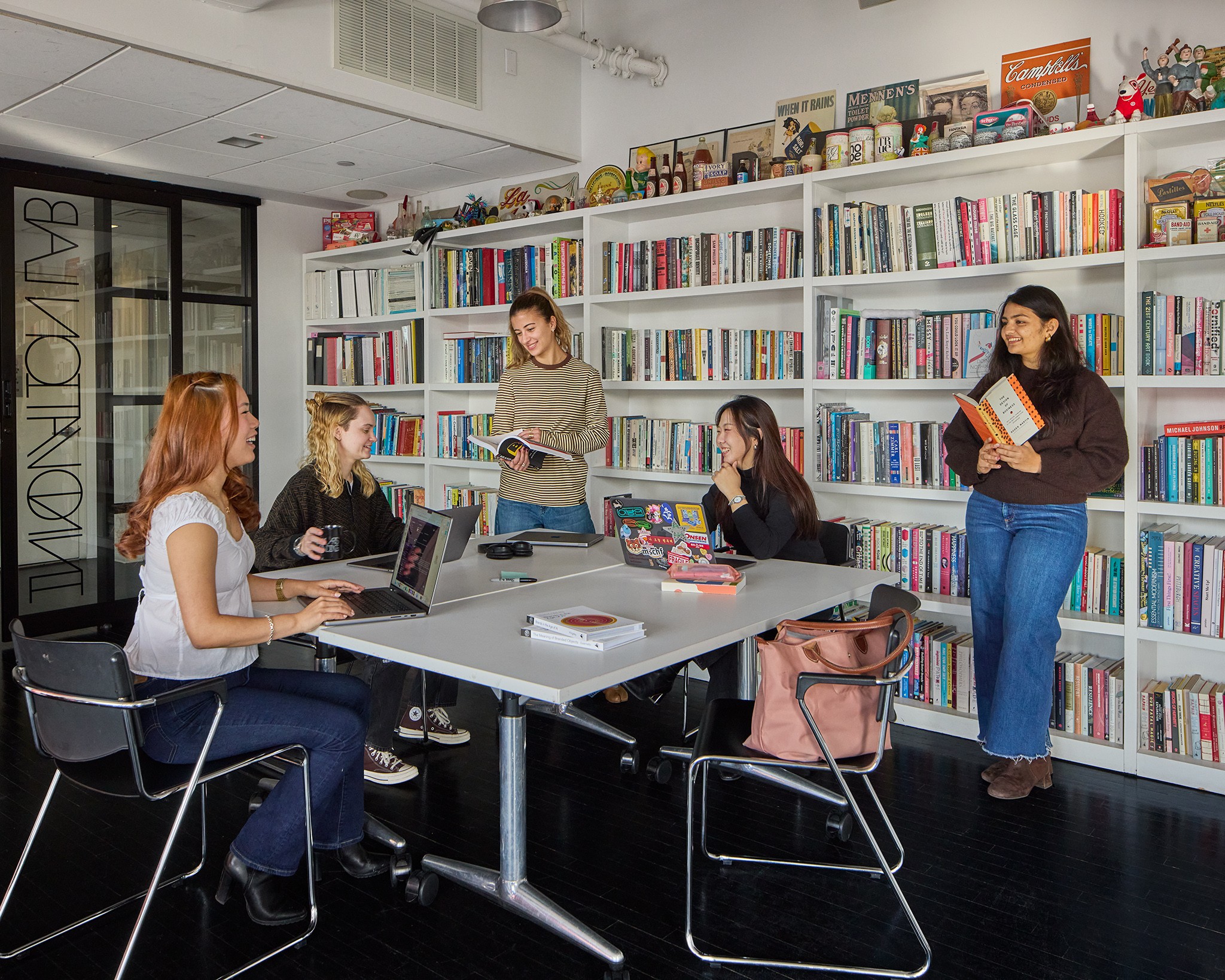 Five women in a library or bookstore, some reading and some using laptops, with shelves filled with books behind them.