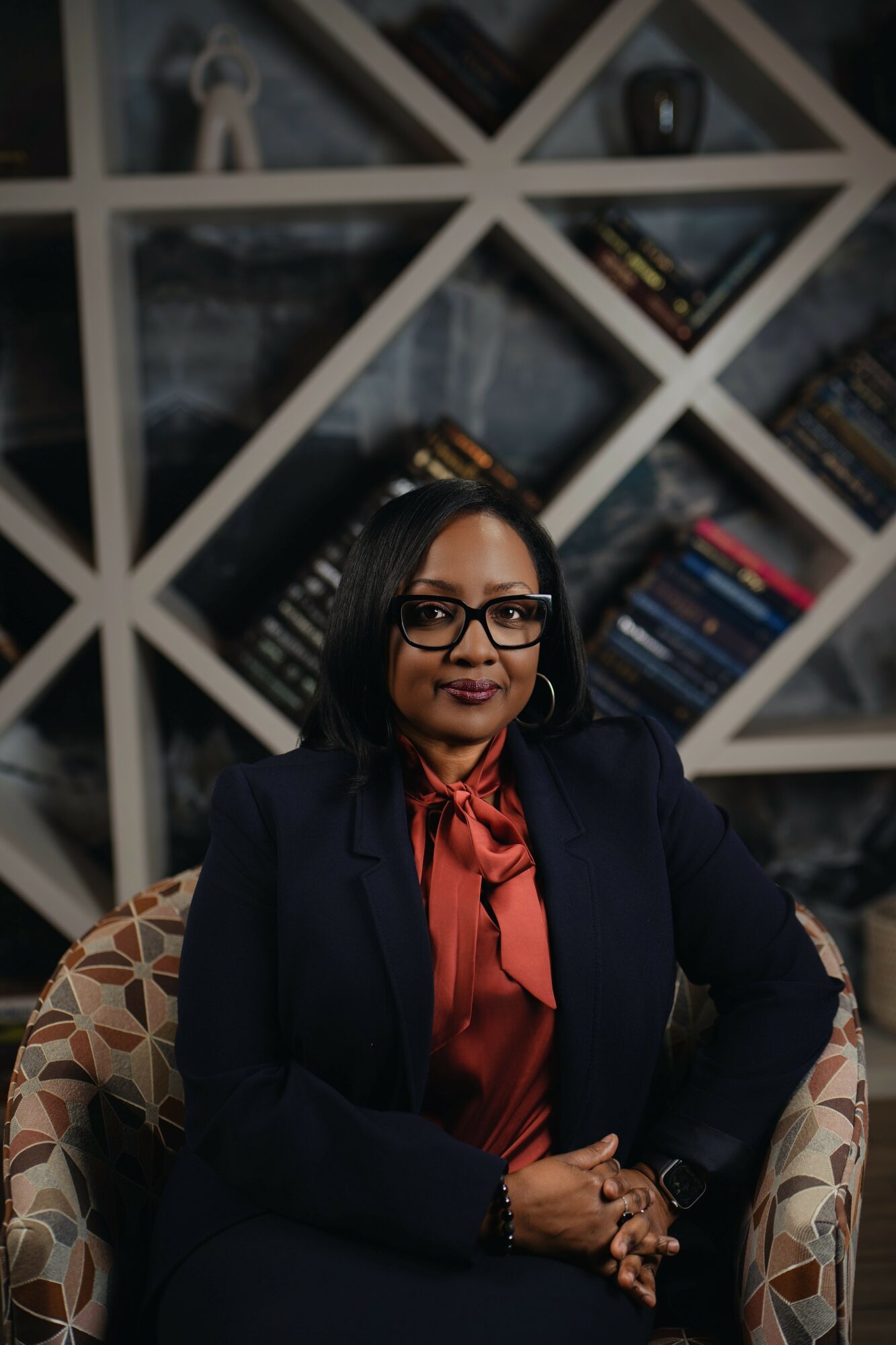 Woman with glasses and dark hair sitting in a patterned chair in front of a geometric bookshelf background.