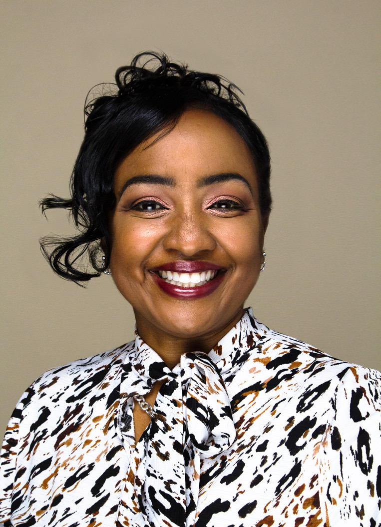Smiling woman with short black hair, wearing a patterned blouse with a bow, against a plain background.