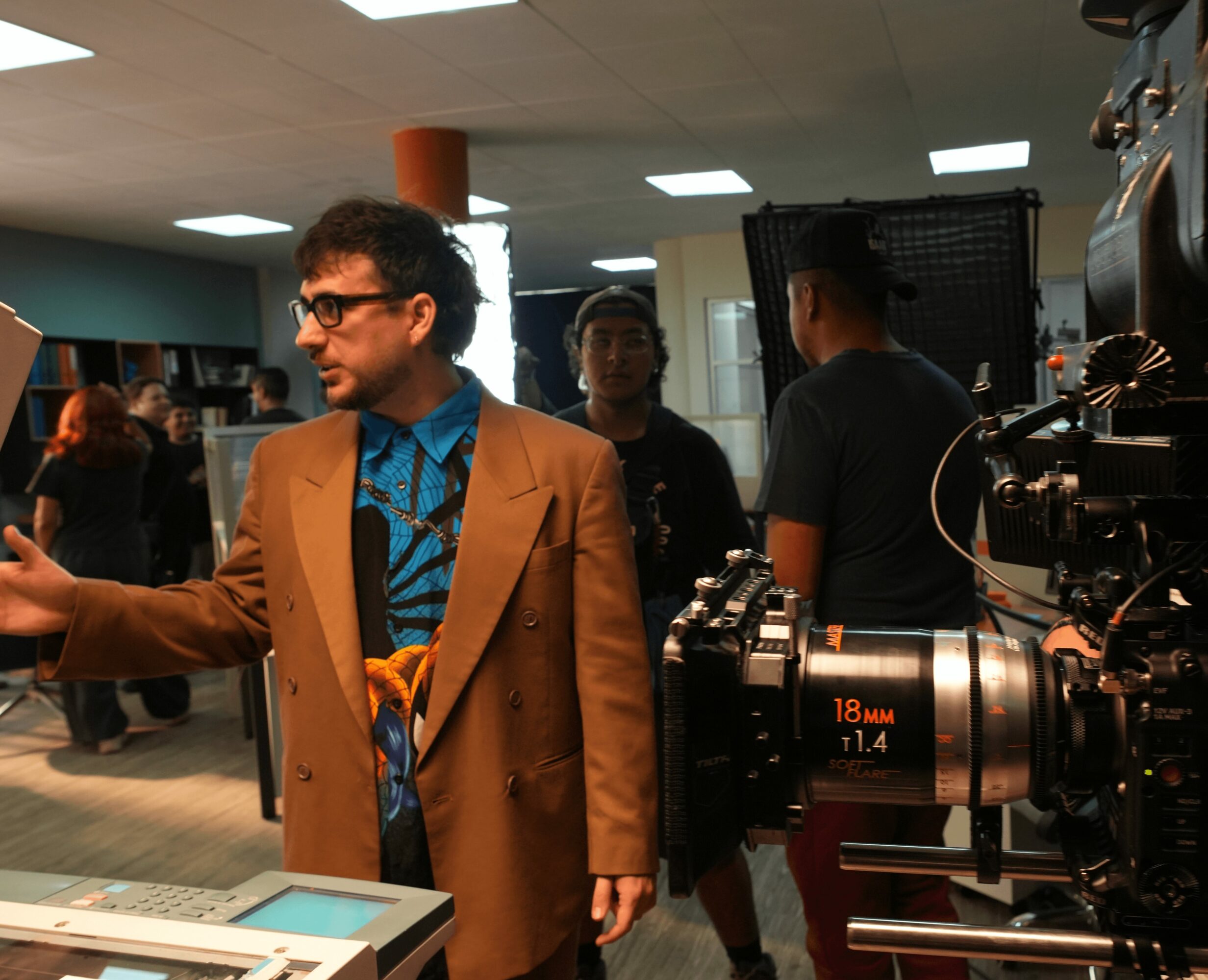 Man in glasses and brown blazer gesturing, surrounded by people and camera equipment indoors.