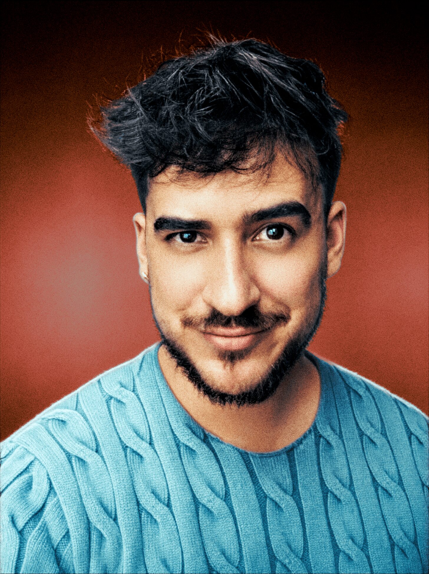 Young man with dark, wavy hair and a beard, smiling, wearing a blue textured shirt, against a reddish background.