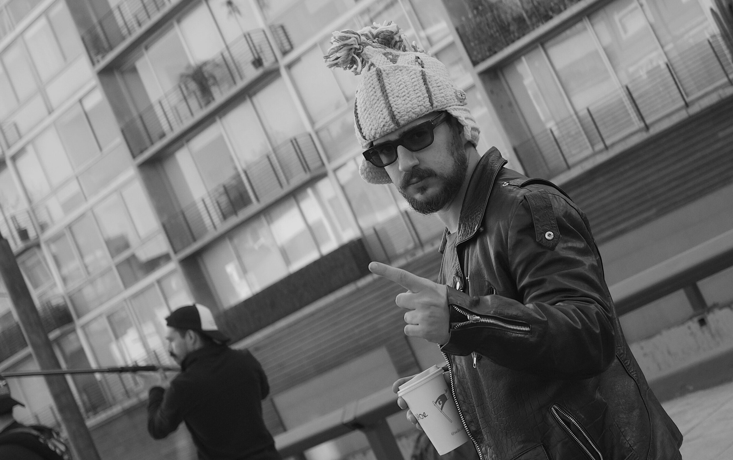 Man wearing sunglasses and leather jacket points at camera, with a person in the background and modern building behind.
