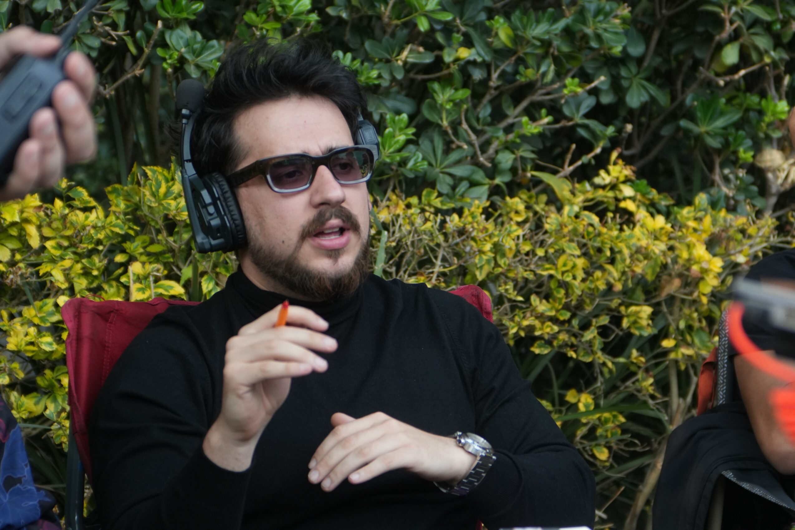 Man with dark hair, beard, sunglasses, and headphones, sitting outdoors with green foliage behind him.