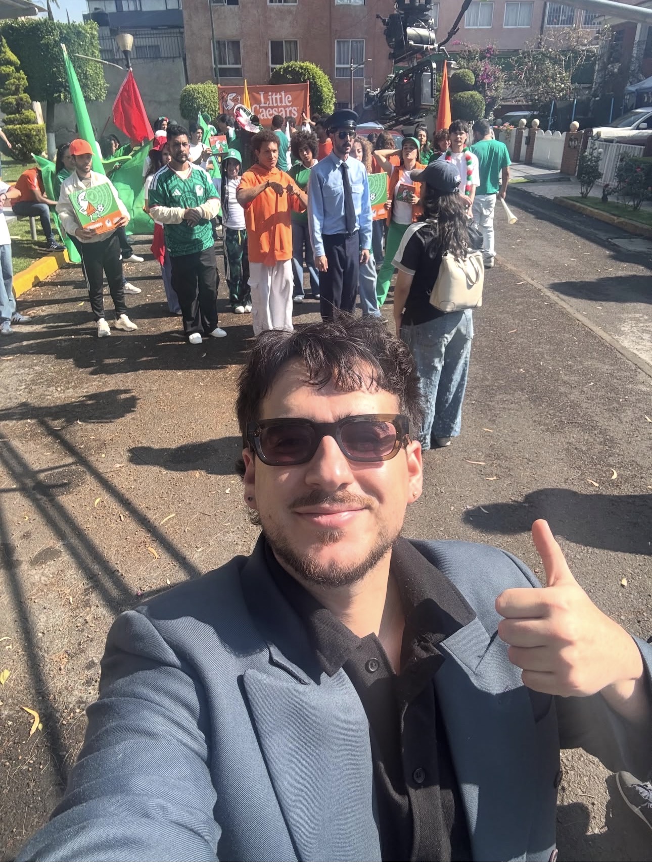 Man taking a selfie outdoors with a group of people holding flags and banners in the background.