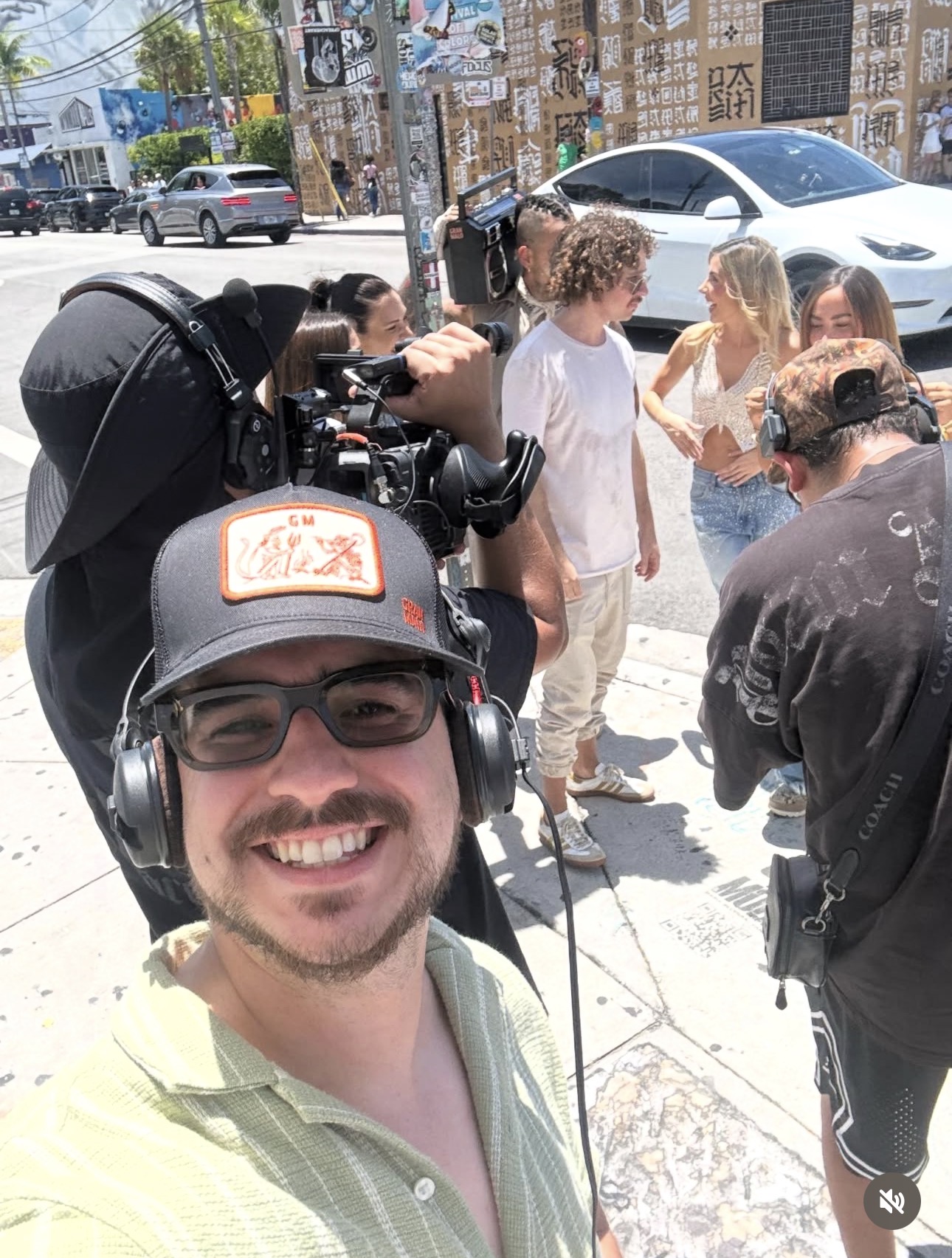 Man wearing glasses and a cap takes a selfie with a group of children and a camera crew on a city sidewalk.