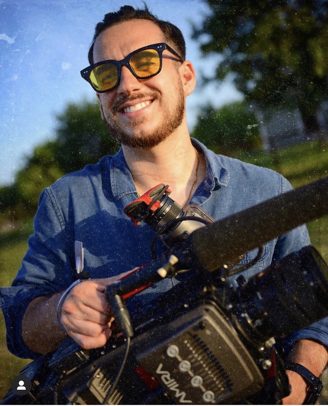 Smiling man with sunglasses holding a camera outdoors, trees and blue sky in background.