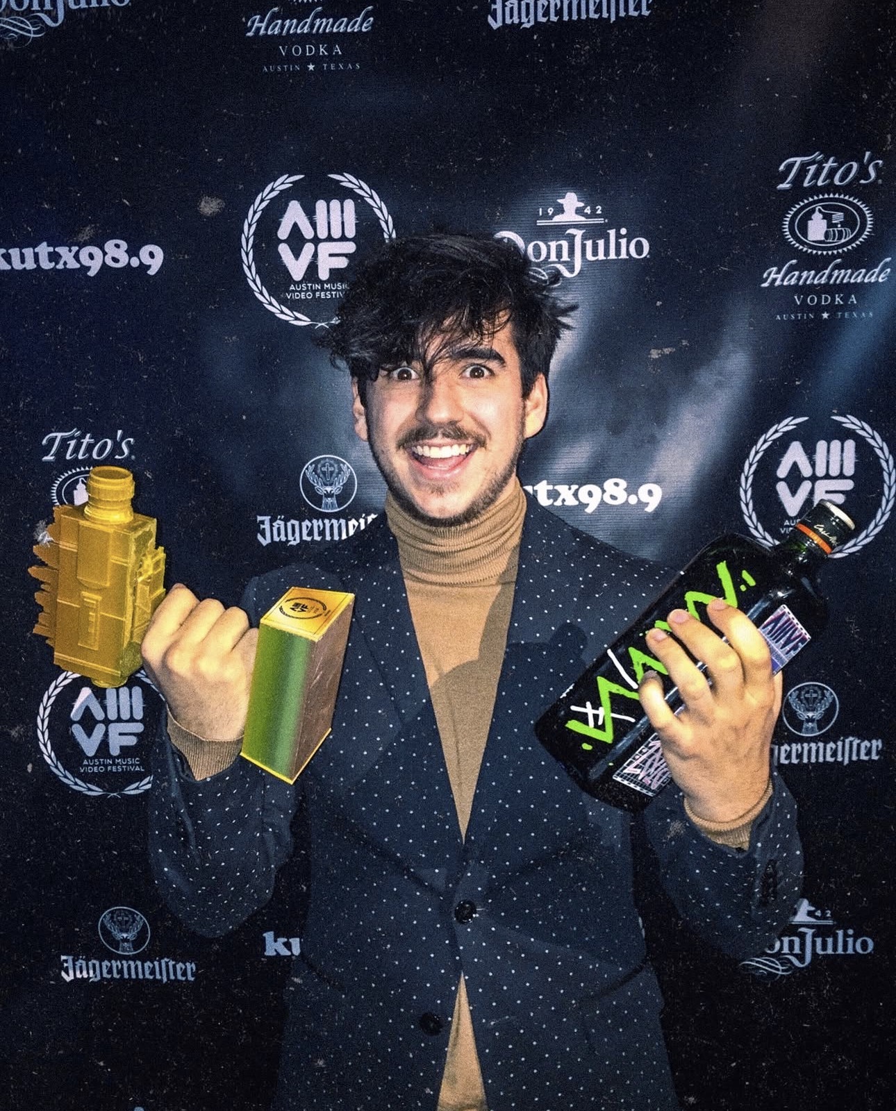 Young man with dark hair and a beard smiling, holding a yellow bottle and a black box, standing in front of a dark backdrop with logos.