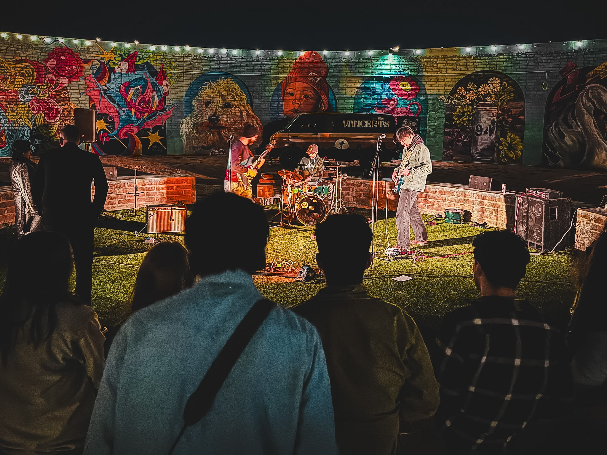 People watching a musical performance outdoors at night with colorful murals in the background.
