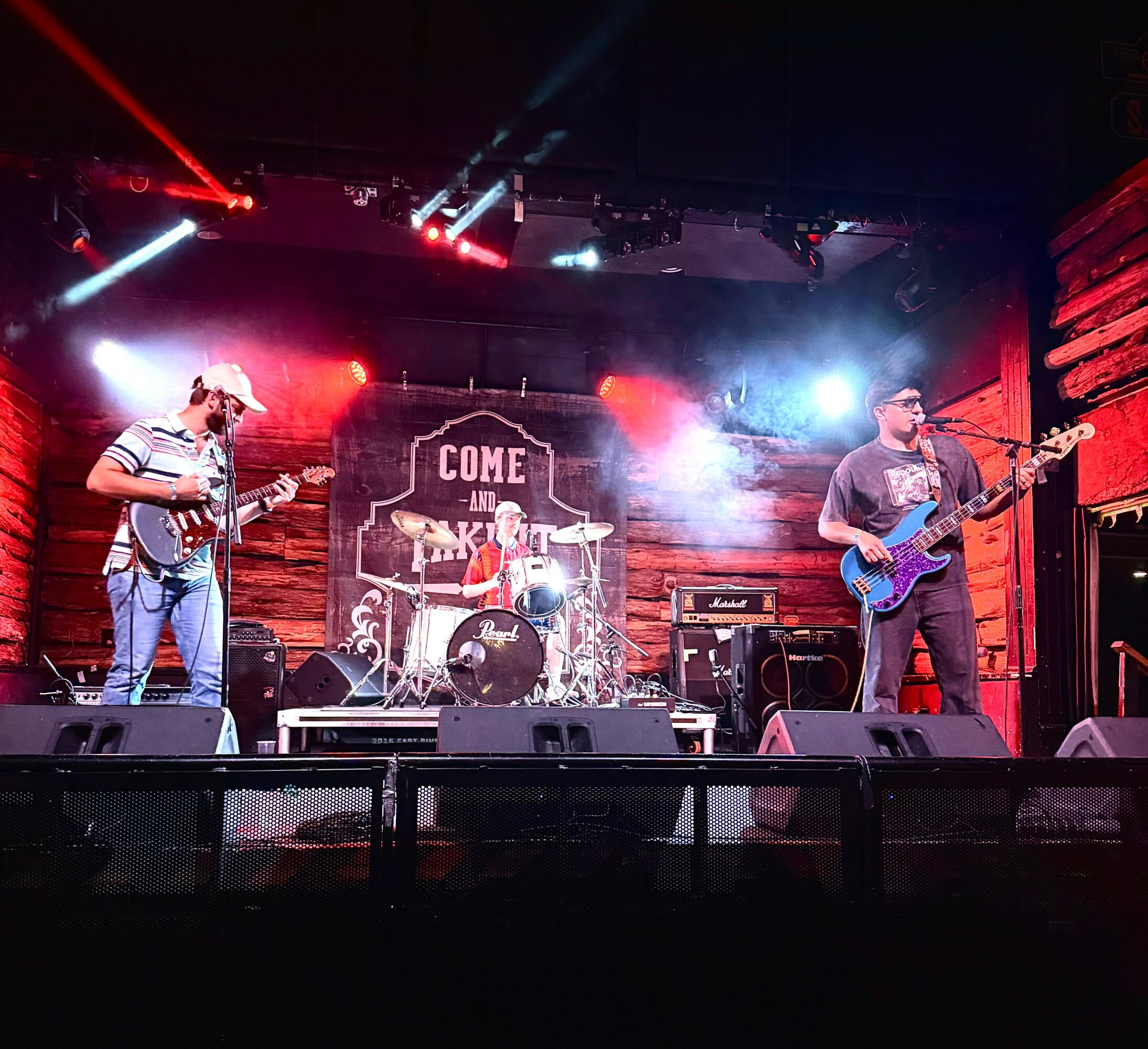 Three musicians perform on stage with guitars and drums, colorful stage lights, and a wooden backdrop with a sign that says 'Come and Take It'.