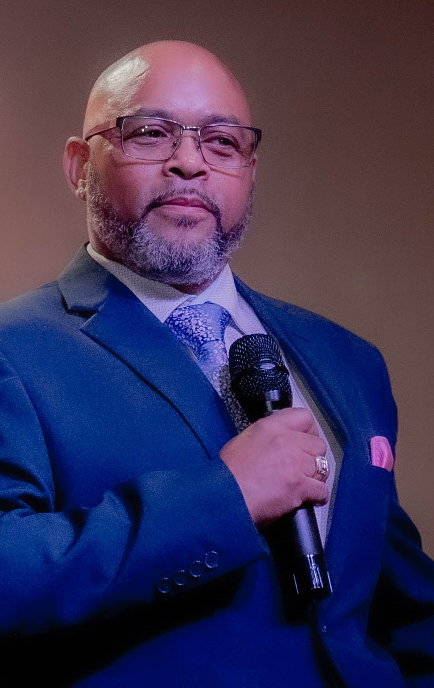 Man with glasses and a beard in a suit holding a microphone, looking to the side.