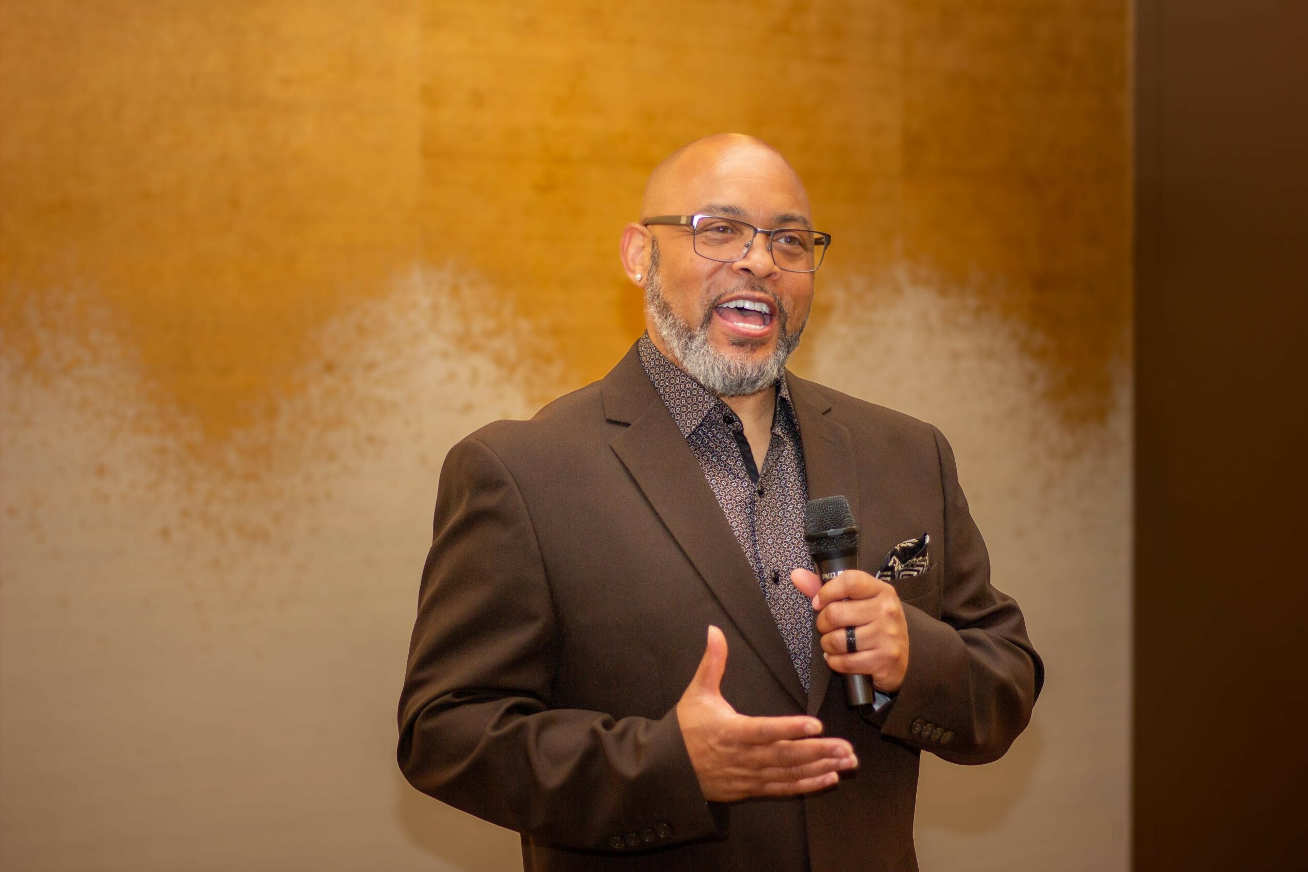 Man with glasses and a beard speaking into a microphone, wearing a dark suit, in front of a yellow and gray background.