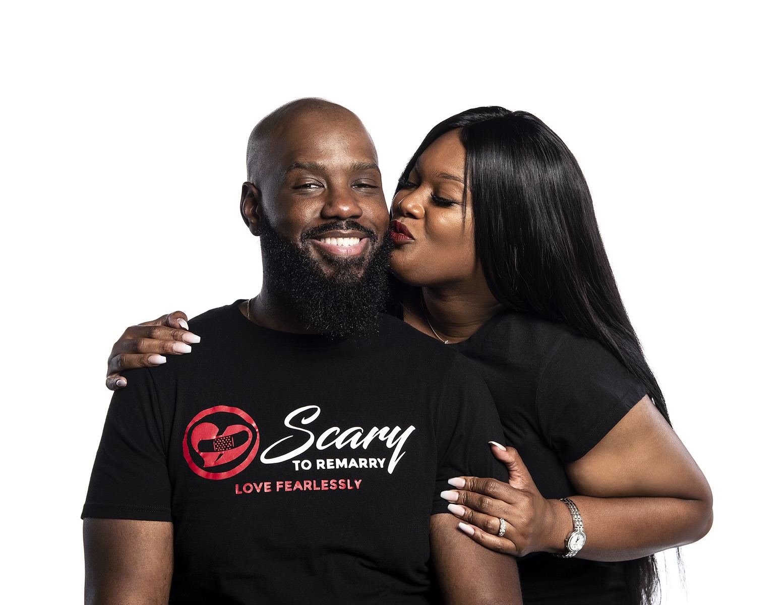A woman kisses a smiling man on the cheek, both wearing black shirts, against a white background.