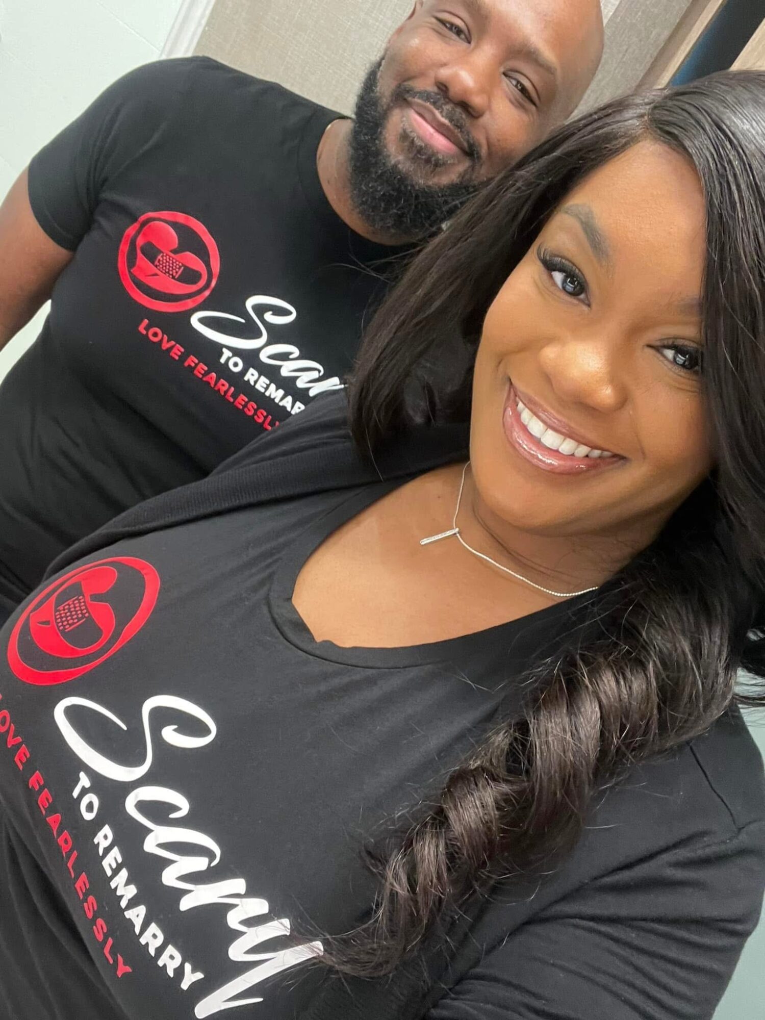 Two people smiling, wearing black shirts with red and white logos and text, close-up selfie, background is plain.