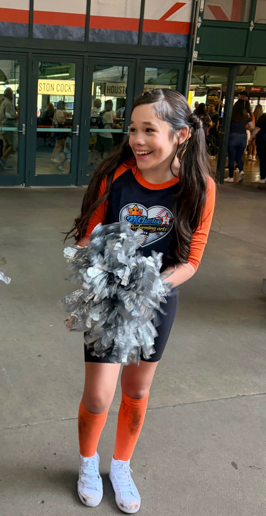 Young girl with pigtails holding pom-poms, smiling, wearing a cheerleading outfit with orange sleeves and socks, and white shoes.