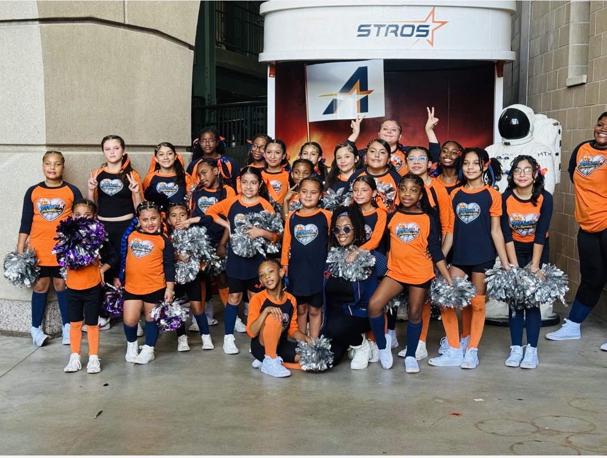 Group of children in cheerleading uniforms holding pom-poms, standing outdoors in front of a stage with a robot figure.
