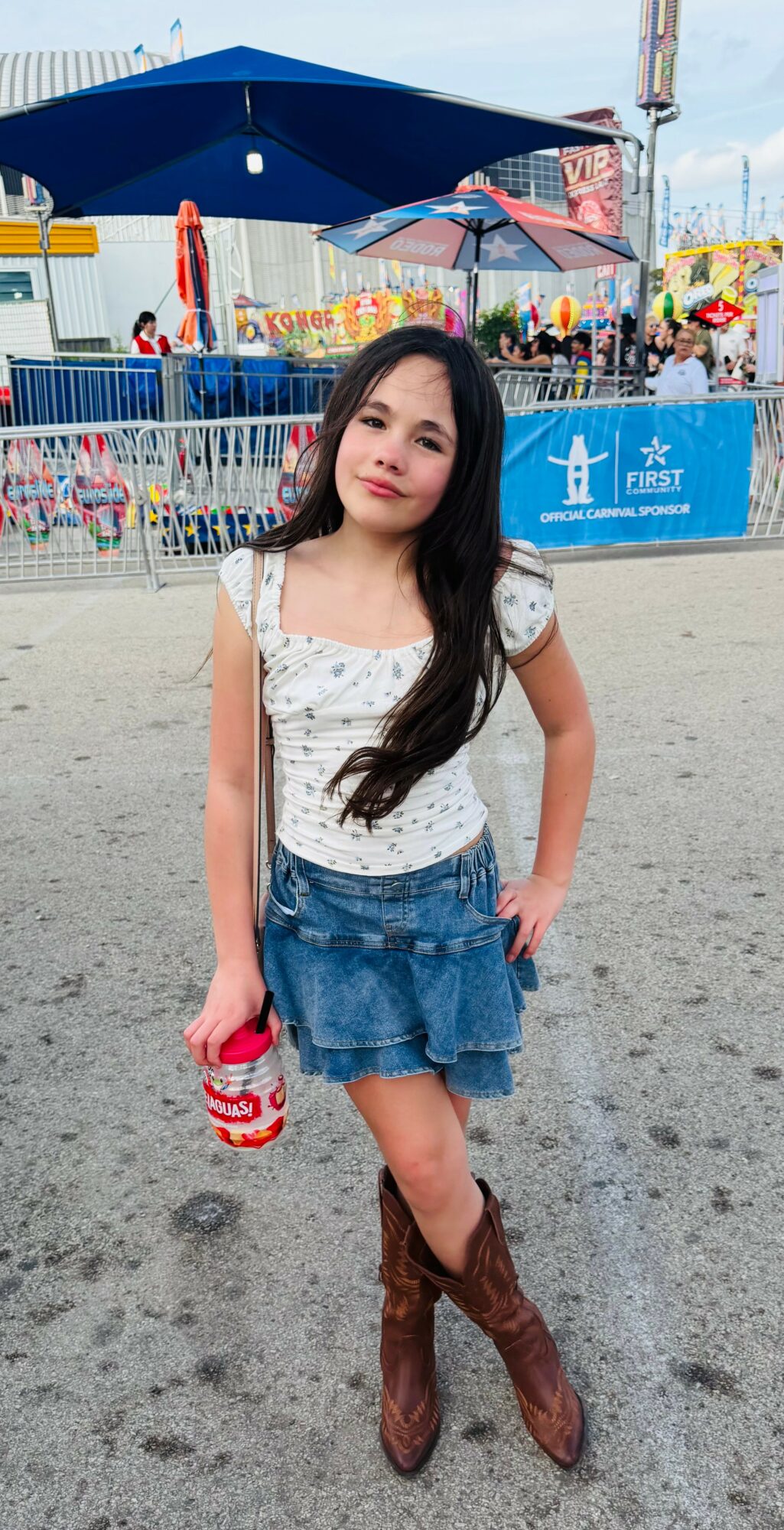 Young girl with long dark hair standing on sandy ground at amusement park, holding a red and white striped cup.