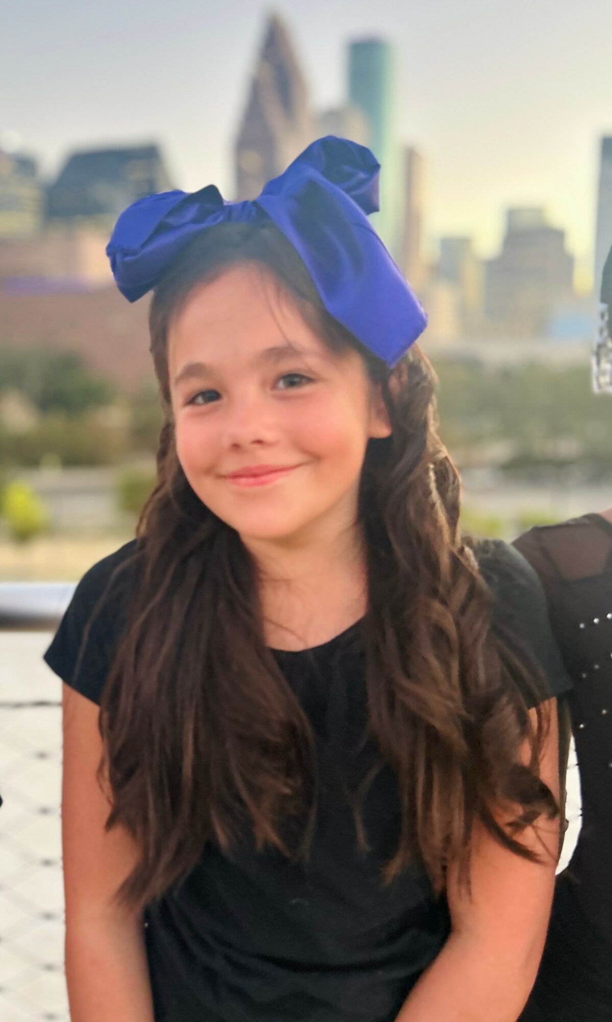 Young girl with long hair and a large blue bow on her head, smiling outdoors with city skyline in background.