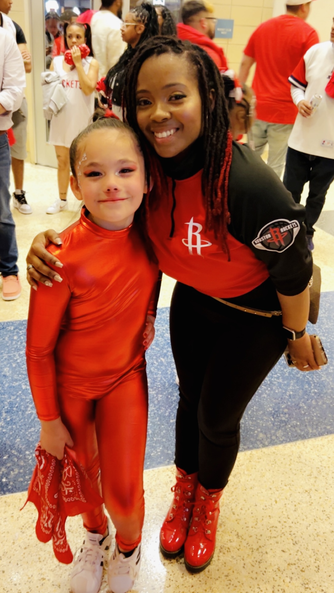 Two smiling girls in red outfits pose together at an indoor event, with other people in the background.