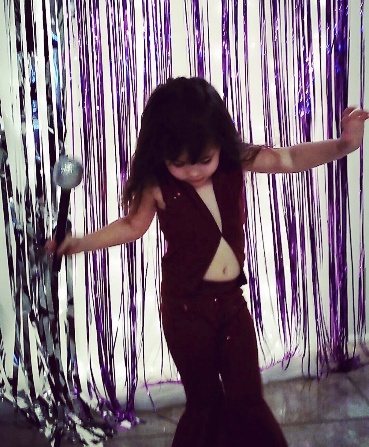 Child with dark hair in a black outfit, standing among purple and silver streamers, holding a microphone, looking down.