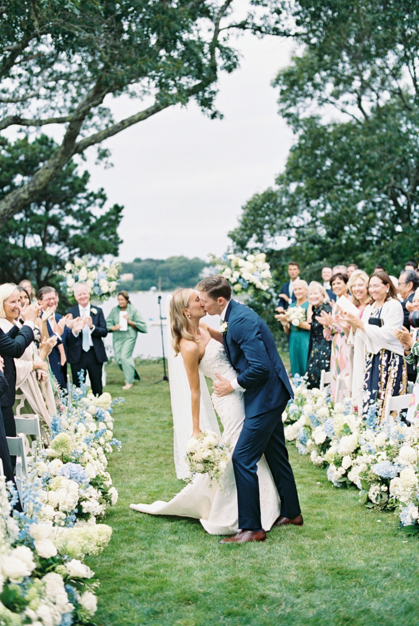 Bride and groom kiss in a garden aisle surrounded by guests and floral arrangements, trees in the background.