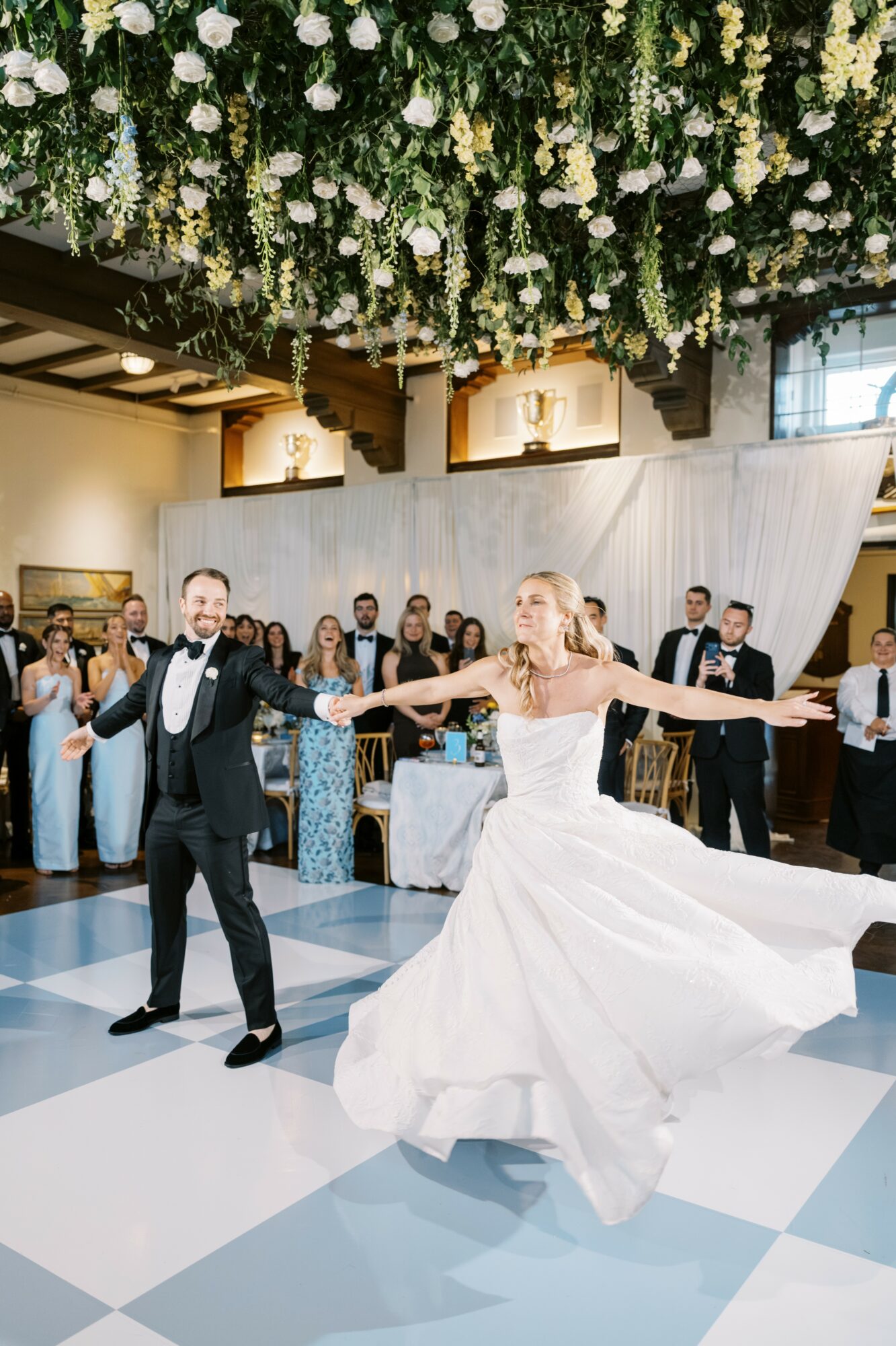 Bride and groom dancing in a decorated wedding venue with guests watching, floral ceiling, and draped backdrop.
