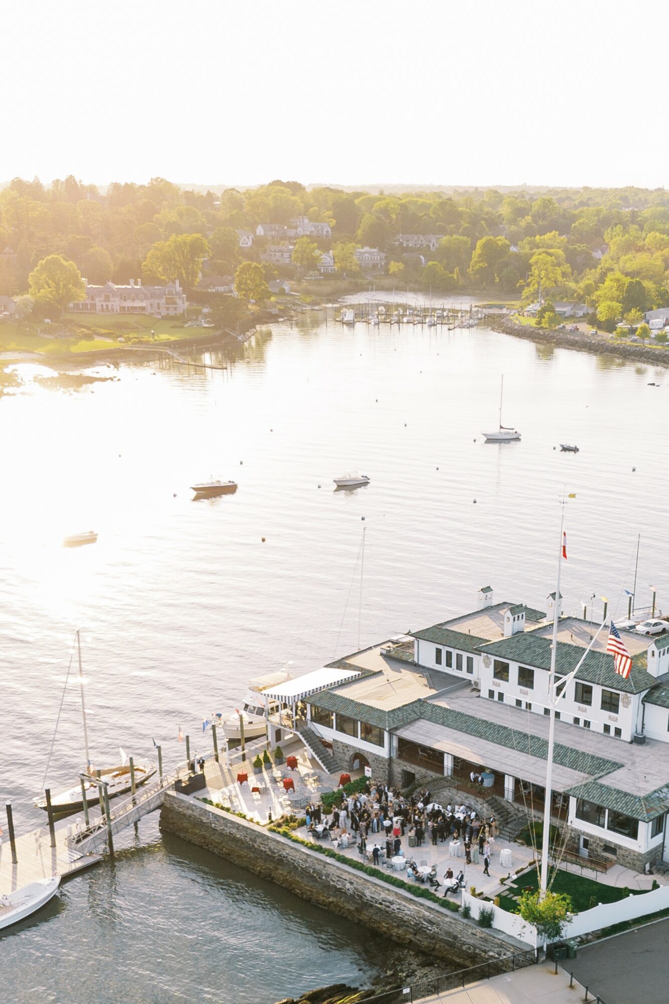 A waterfront restaurant with outdoor seating, boats on the water, and a tree-lined shoreline in the background.