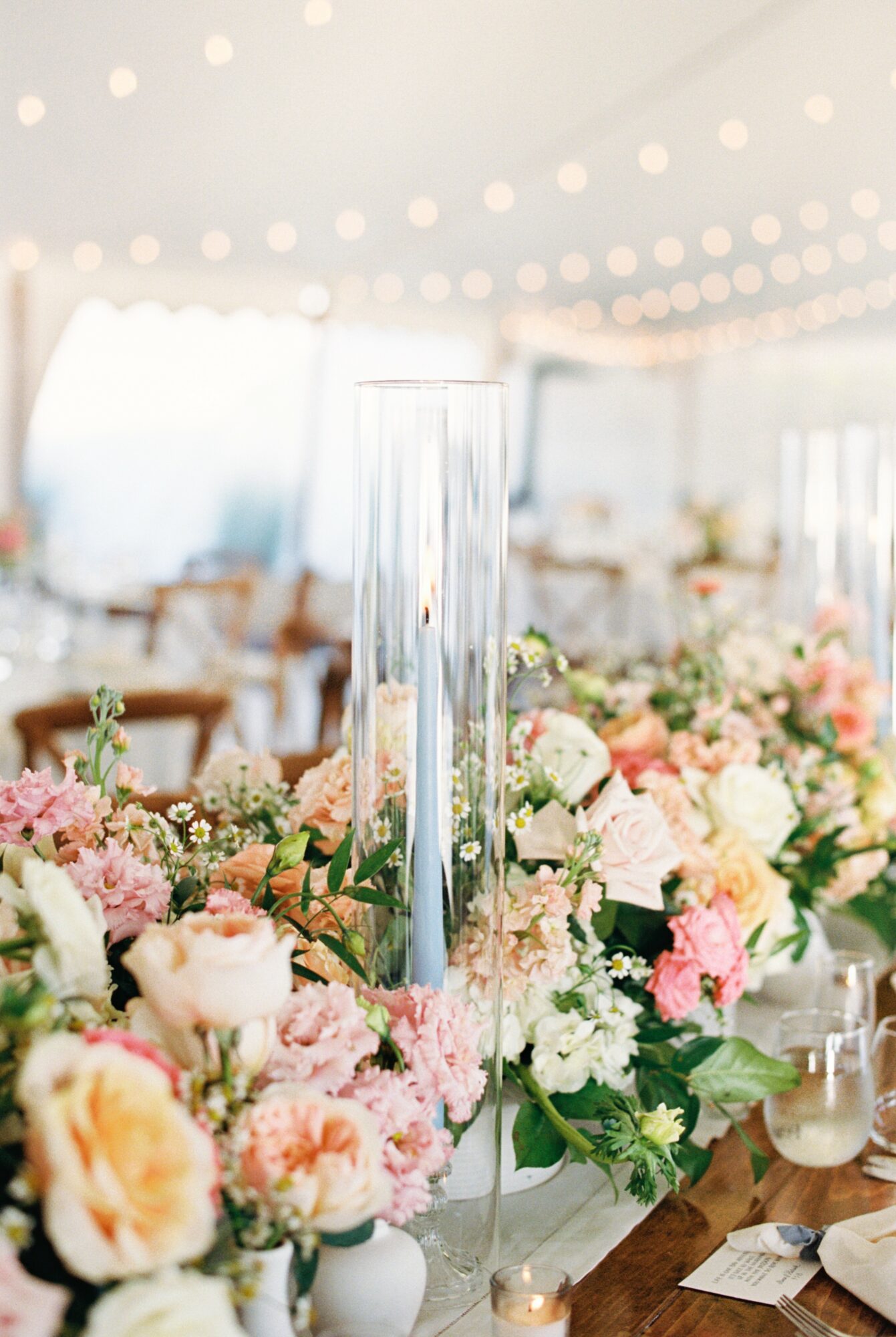 Tall glass vase with a single flower stem among pink and white flowers on a decorated table, blurred background with string lights