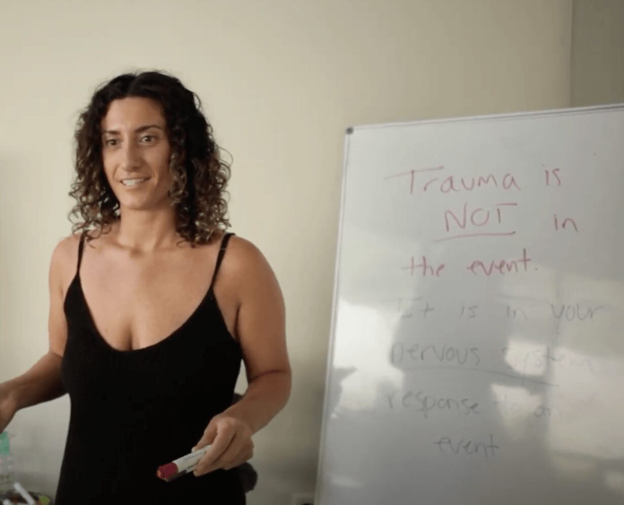 Woman with curly hair in a black top standing next to a whiteboard with handwritten text, holding a marker.