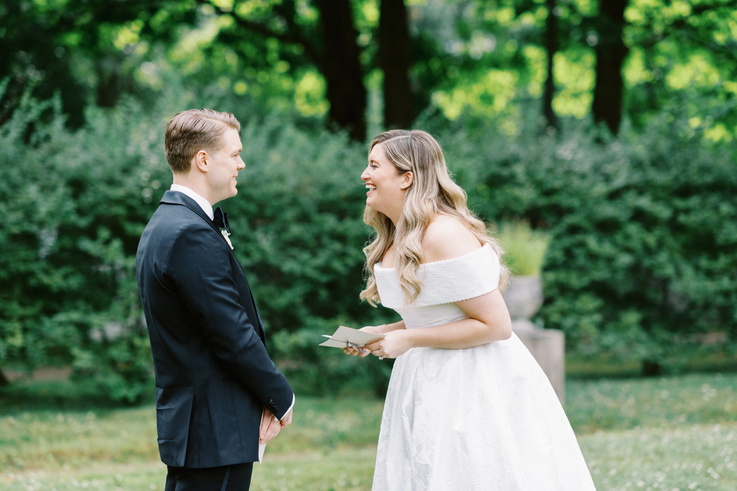 A woman in a white dress and a man in a suit face each other outdoors, smiling, with trees in the background.