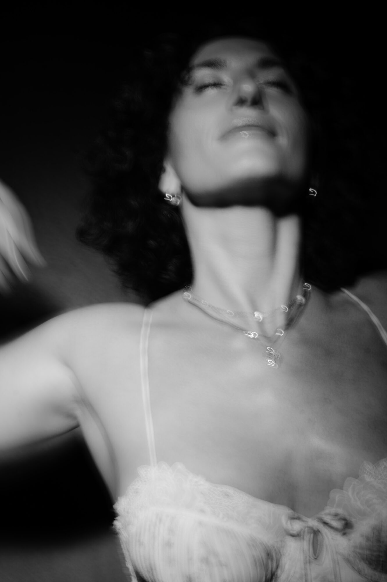 Woman with curly hair and closed eyes, wearing a spaghetti strap top and jewelry, looking upward.