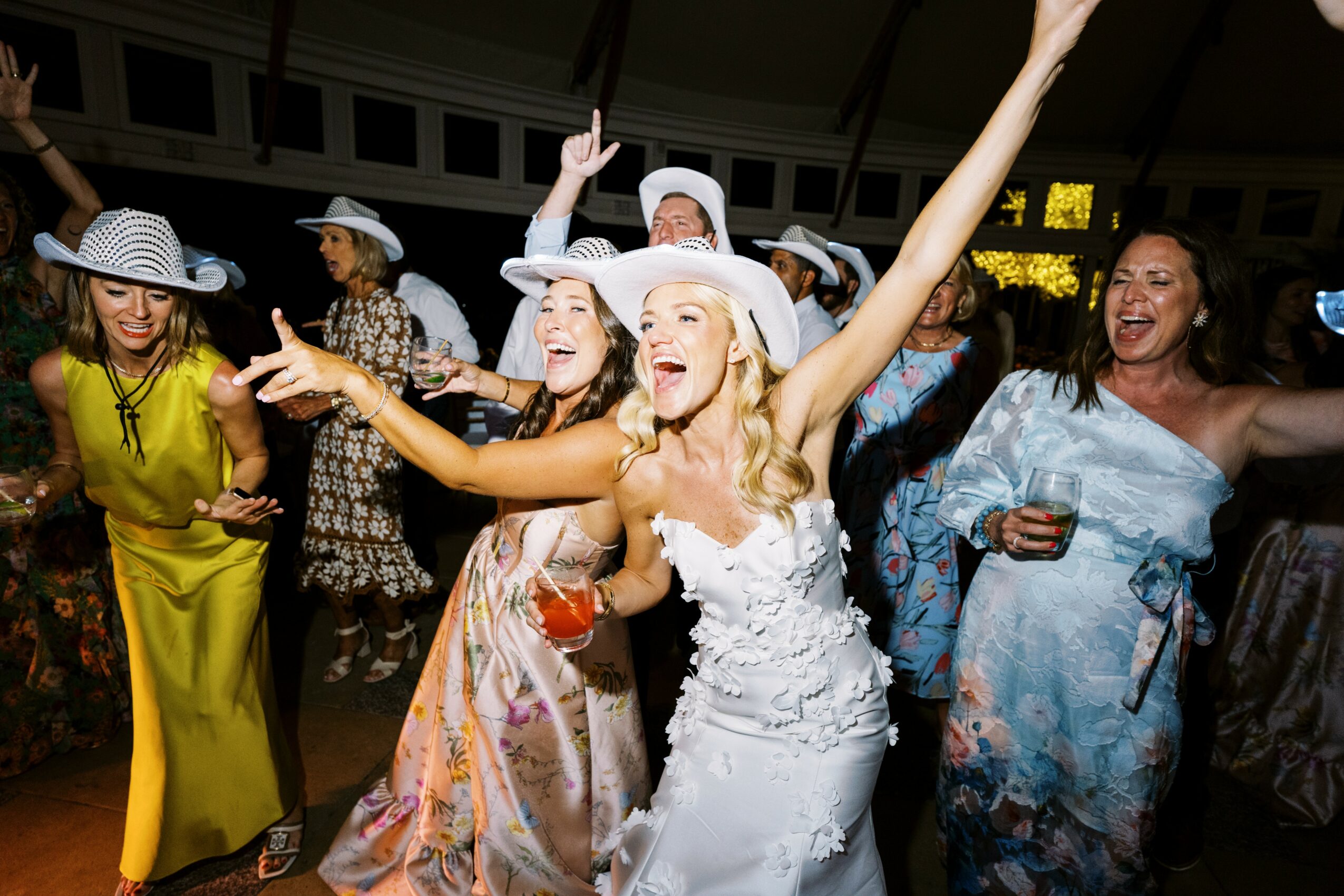 Group of women celebrating, smiling, and dancing at a party, some holding drinks, wearing summer dresses and hats.