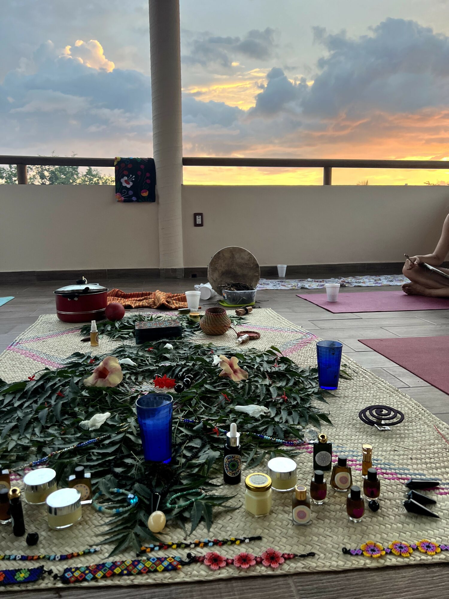 Table with various items, greenery, candles, and bottles, set outdoors during sunset with a railing and sky in background.