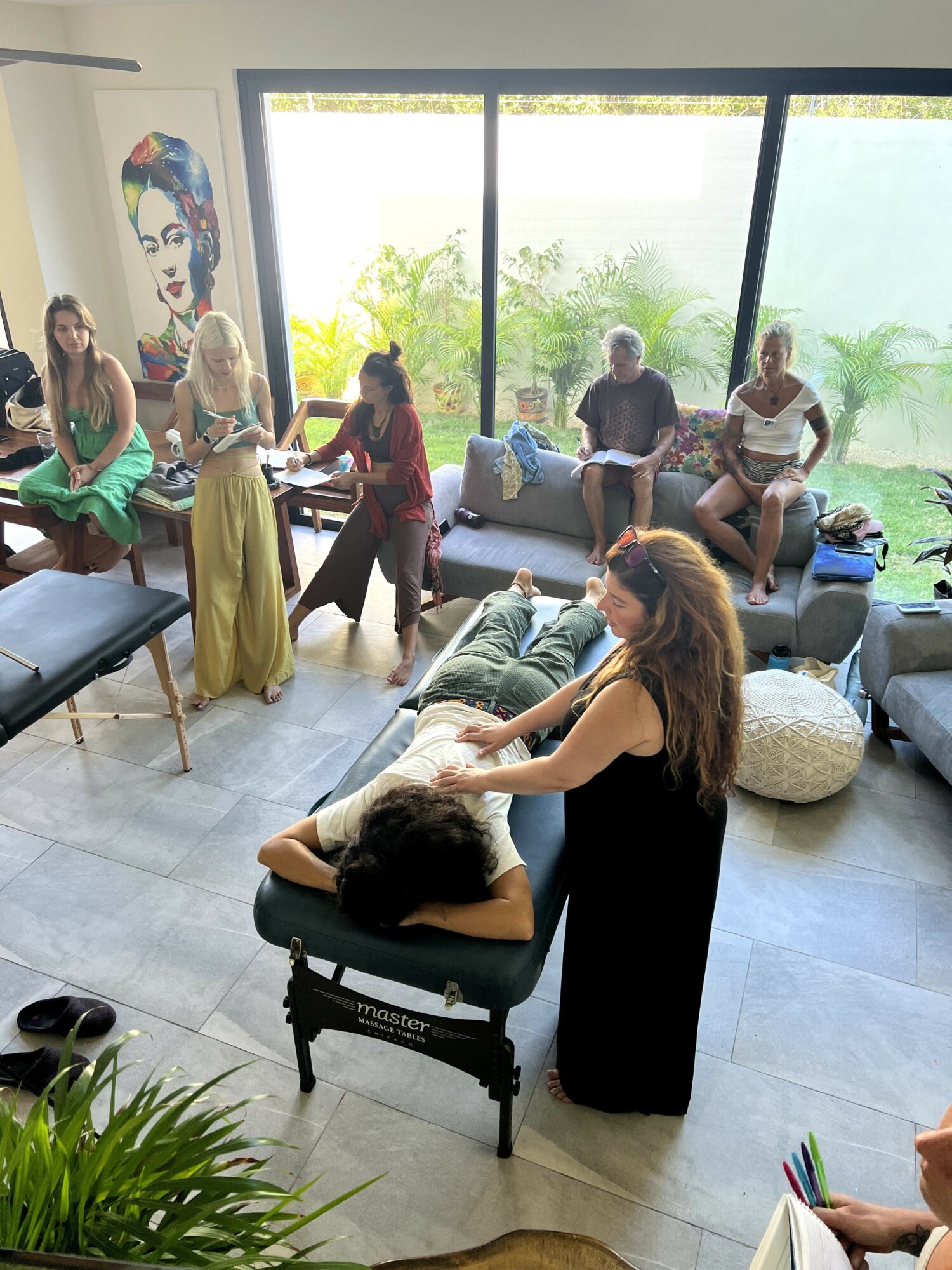 Group of people in a room with large windows, some sitting on sofas, one person receiving a massage, colorful artwork on wall.