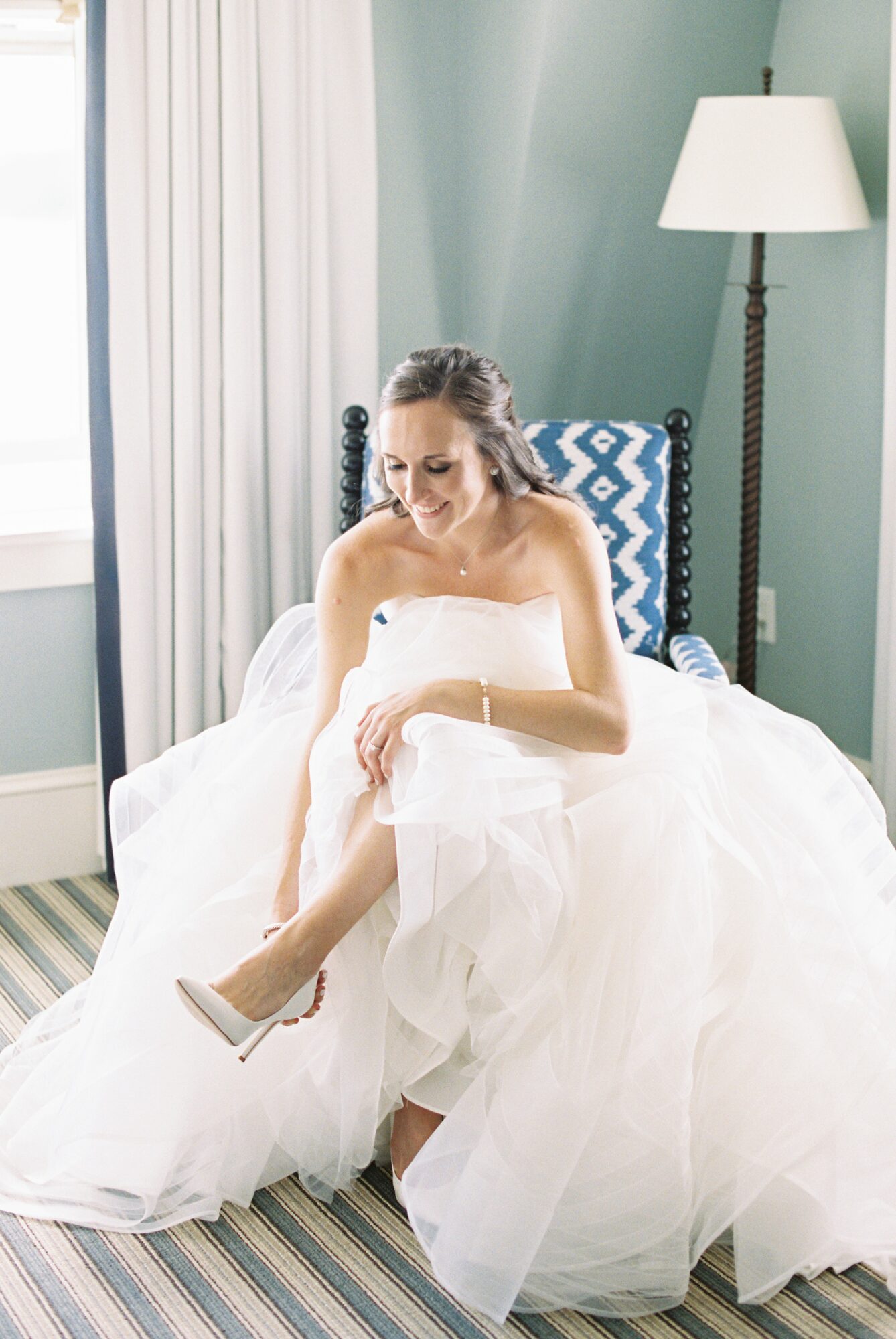 Woman in wedding dress sitting on bed, adjusting her shoe, in a room with a lamp and patterned pillow.