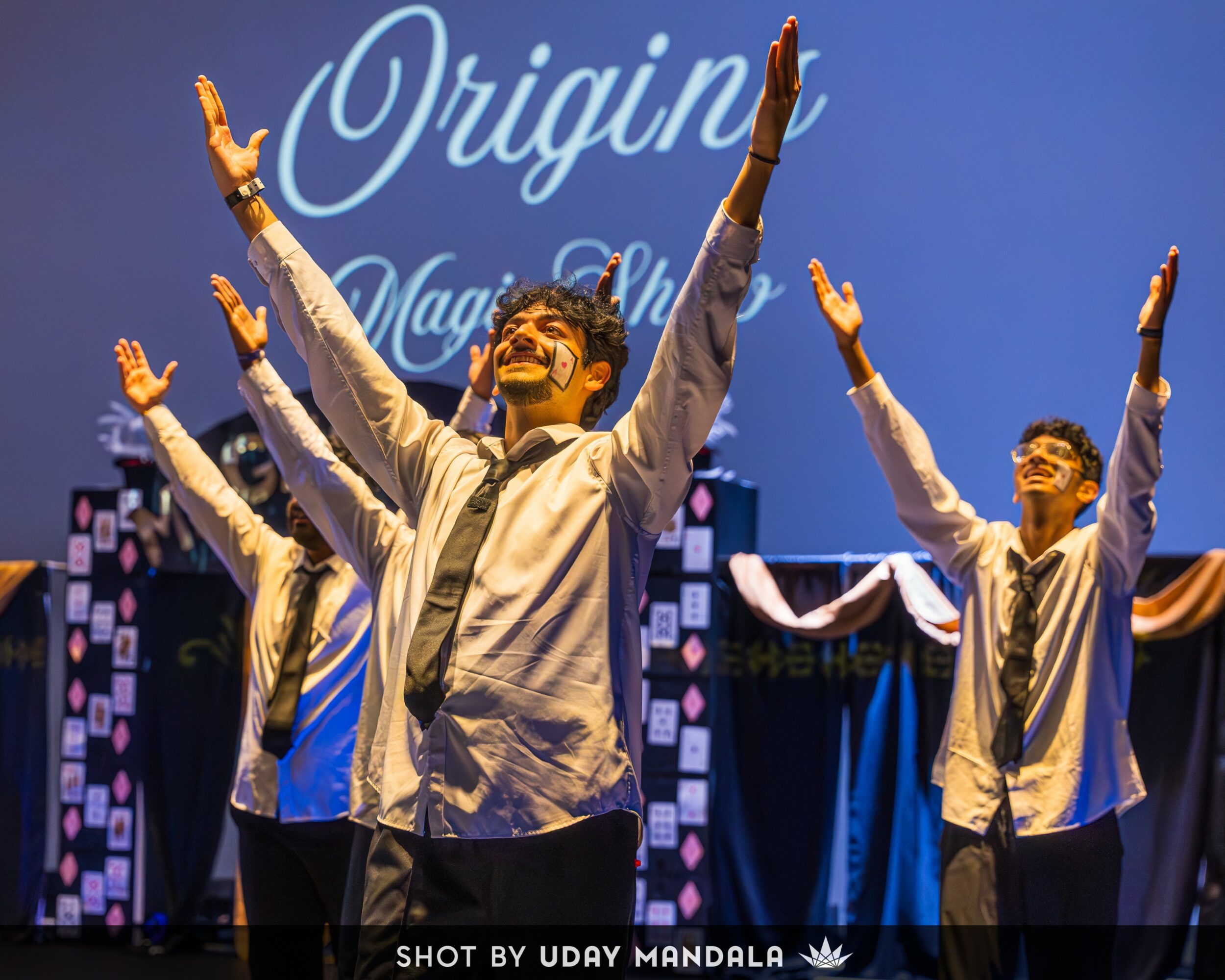 Group of young men on stage with arms raised, smiling, with a blue background and the word 'Origin' projected behind them.
