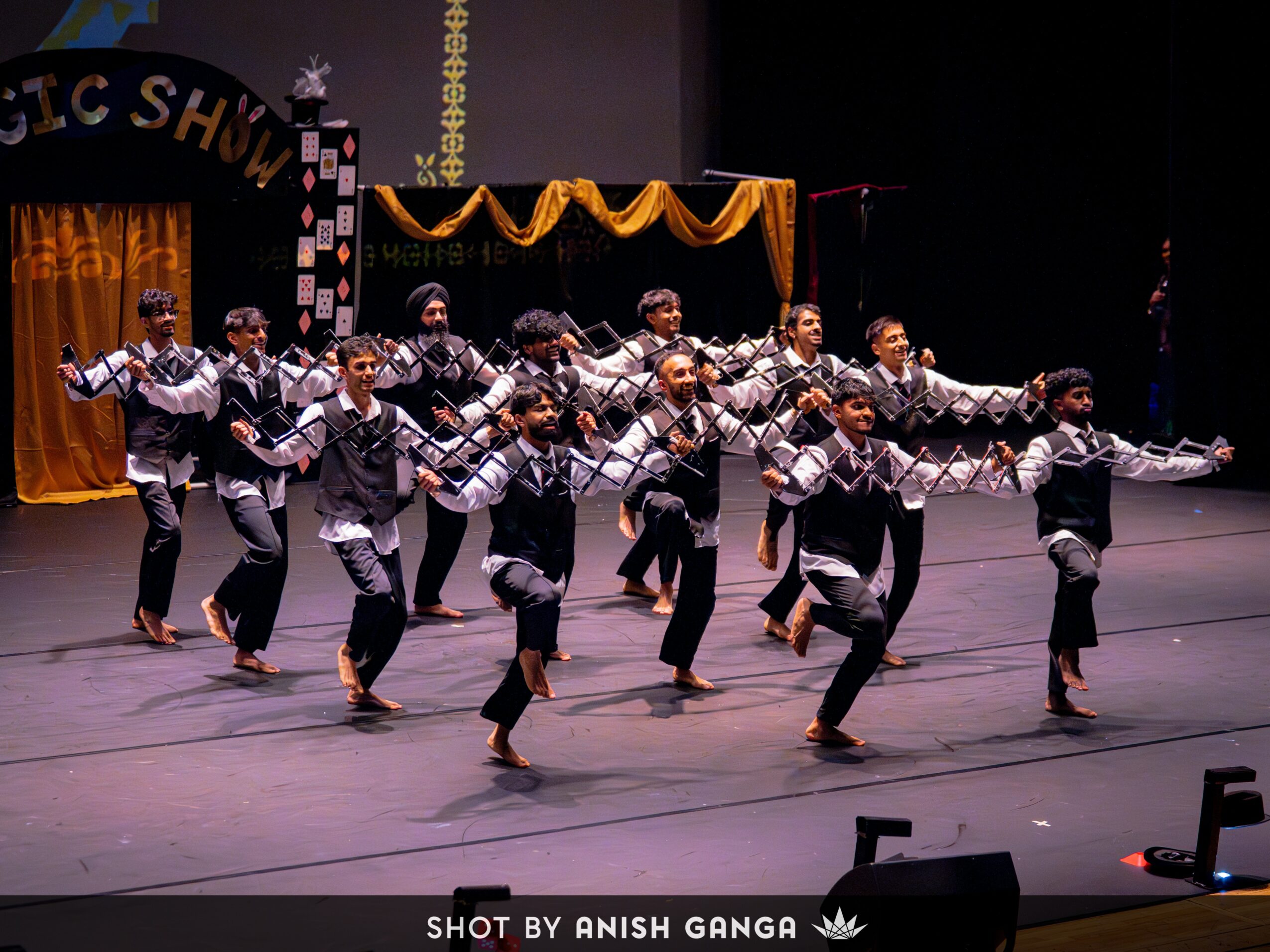 Group of performers dancing on stage with arms extended, dressed in coordinated costumes, stage decorated with curtains and banners.