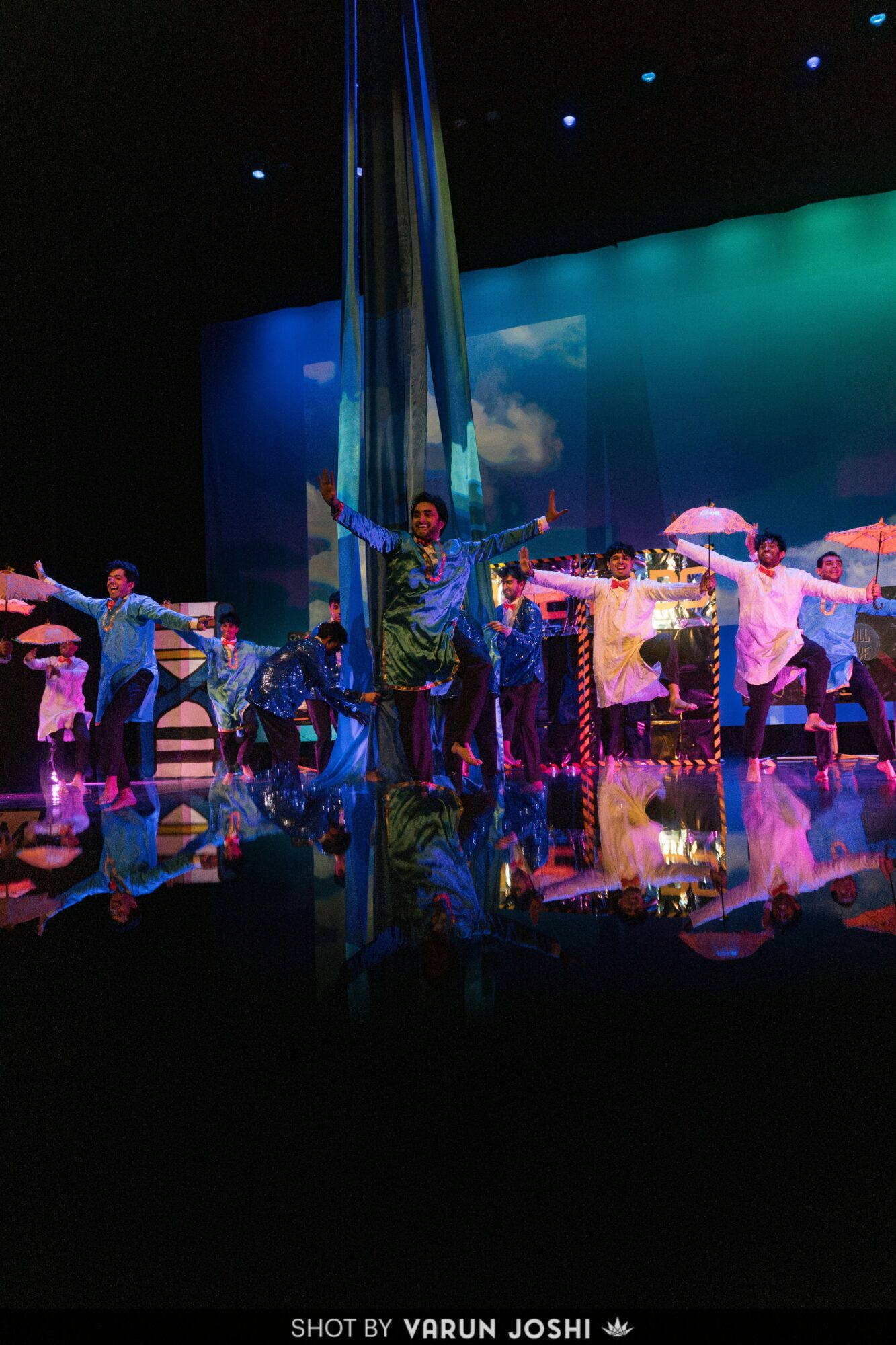 Stage performance with dancers in colorful costumes, set against a bright, scenic backdrop, with a central performer holding an umbrella.
